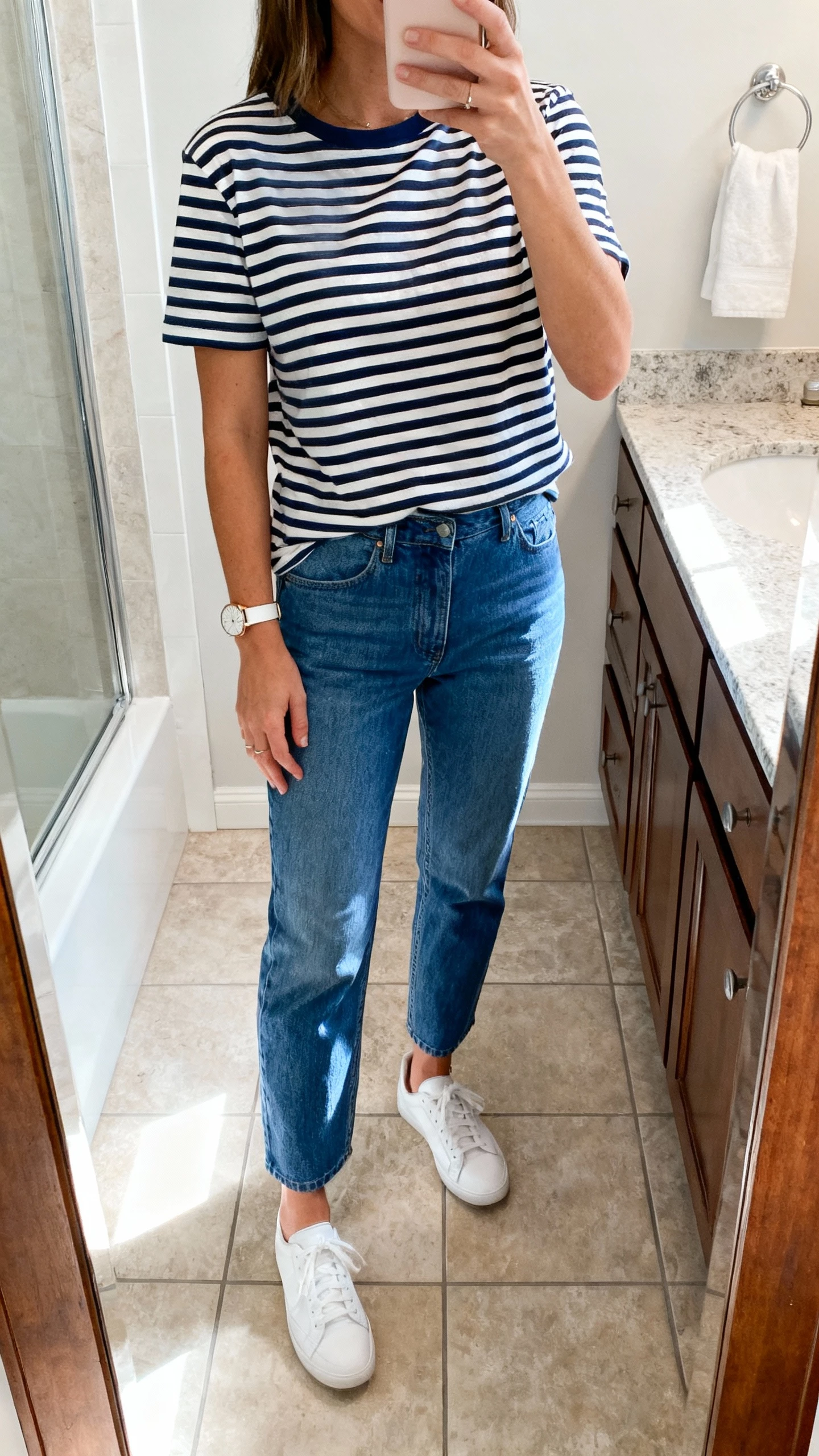 iPhone mirror selfie of a woman wearing a classic navy-and-white Breton striped tee with straight-leg blue jeans, white sneakers, simple watch, face not visible, bathroom mirror, bright natural light, iPhone photo quality.