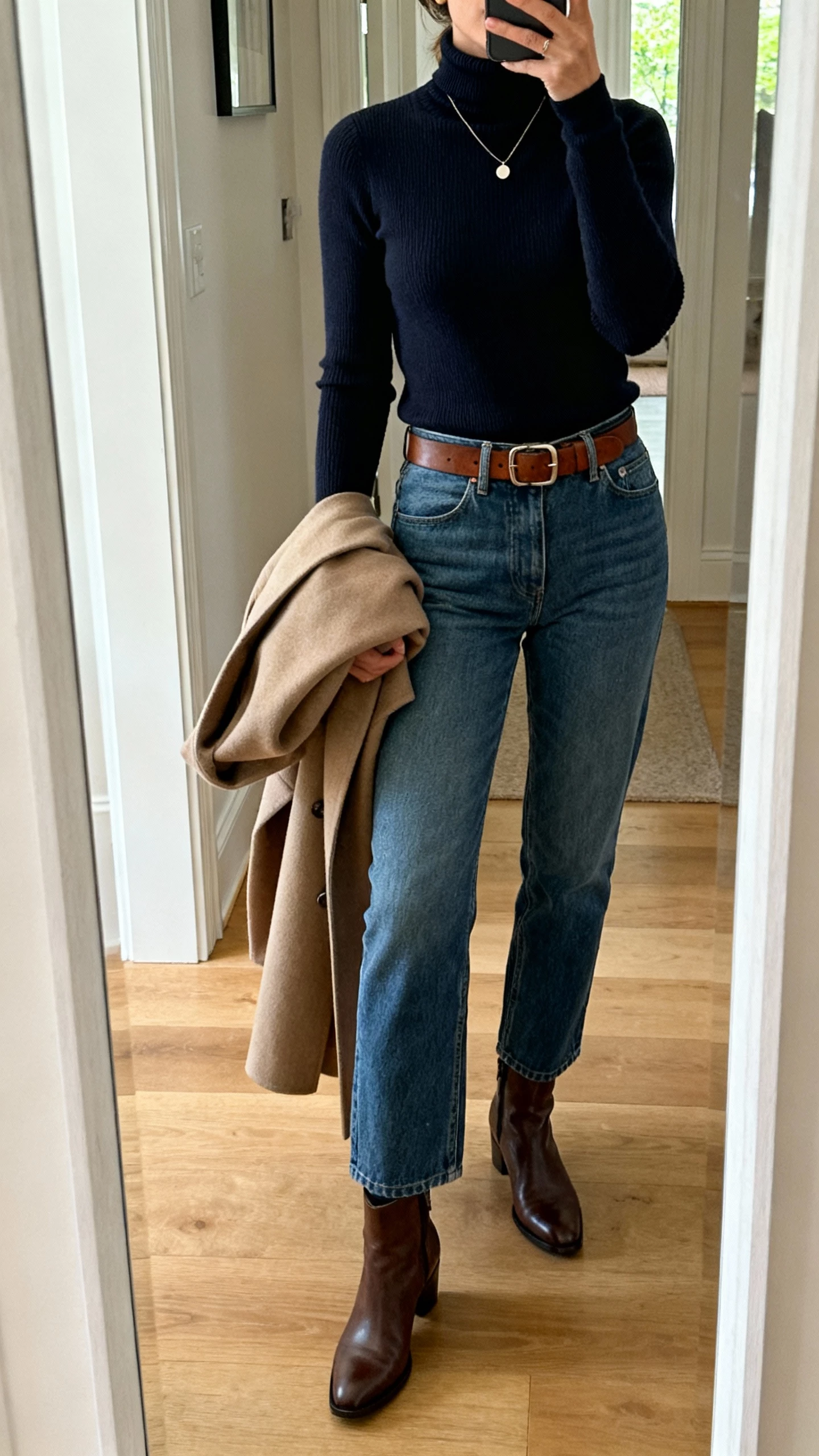 iPhone mirror selfie of a woman wearing a classic turtleneck tucked into straight-leg denim with ankle boots, leather belt, delicate necklace, structured coat draped over arm, face not visible, entryway mirror, natural daylight, natural iPhone photo quality.