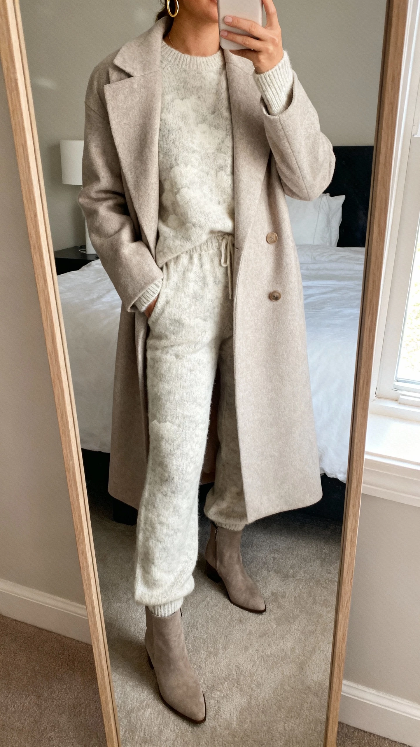 iPhone mirror selfie of a woman wearing a cloud-soft knit set (matching cozy sweater and knit pants) with a polished long coat, minimal gold hoops, ankle boots, face not visible, cropped at neck, bedroom mirror with morning window light, natural iPhone photo quality.