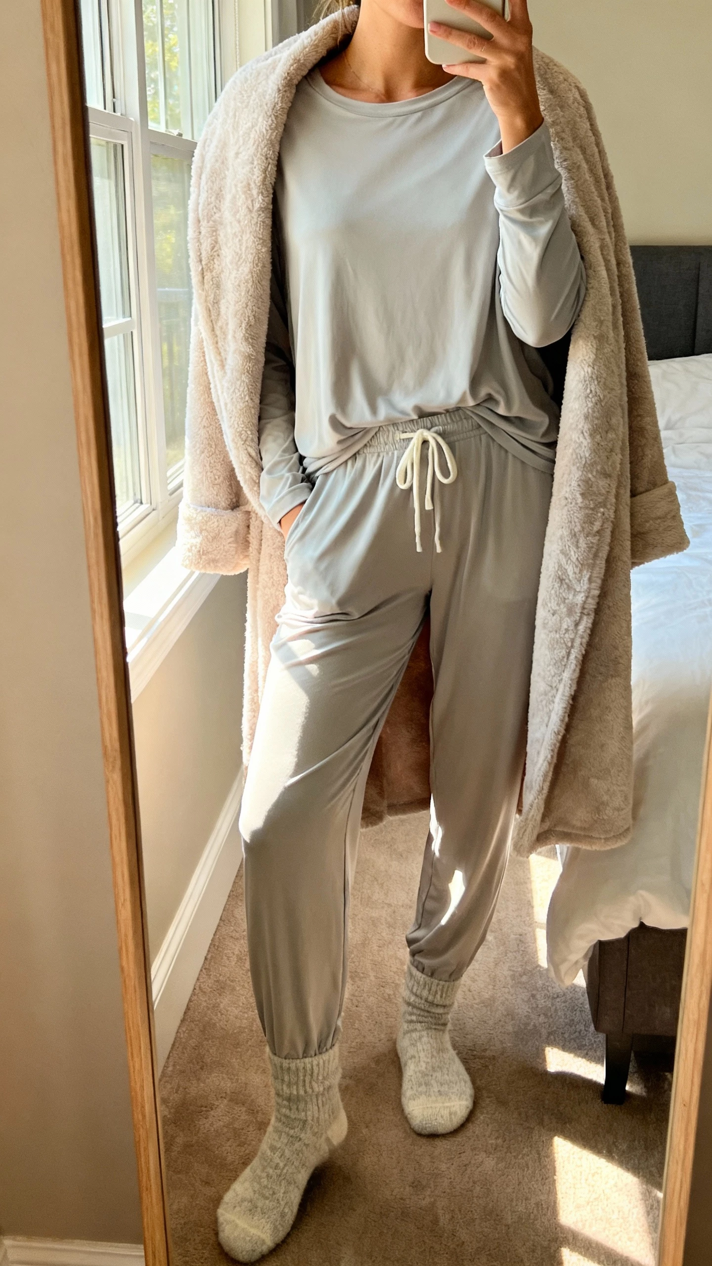 iPhone mirror selfie of a woman wearing a cloud-soft modal pajama set (long-sleeve top and matching drawstring pants) with cozy socks and a plush robe draped over shoulders, face not visible, cropped at neck, bedroom mirror with warm morning window light, natural iPhone photo quality.