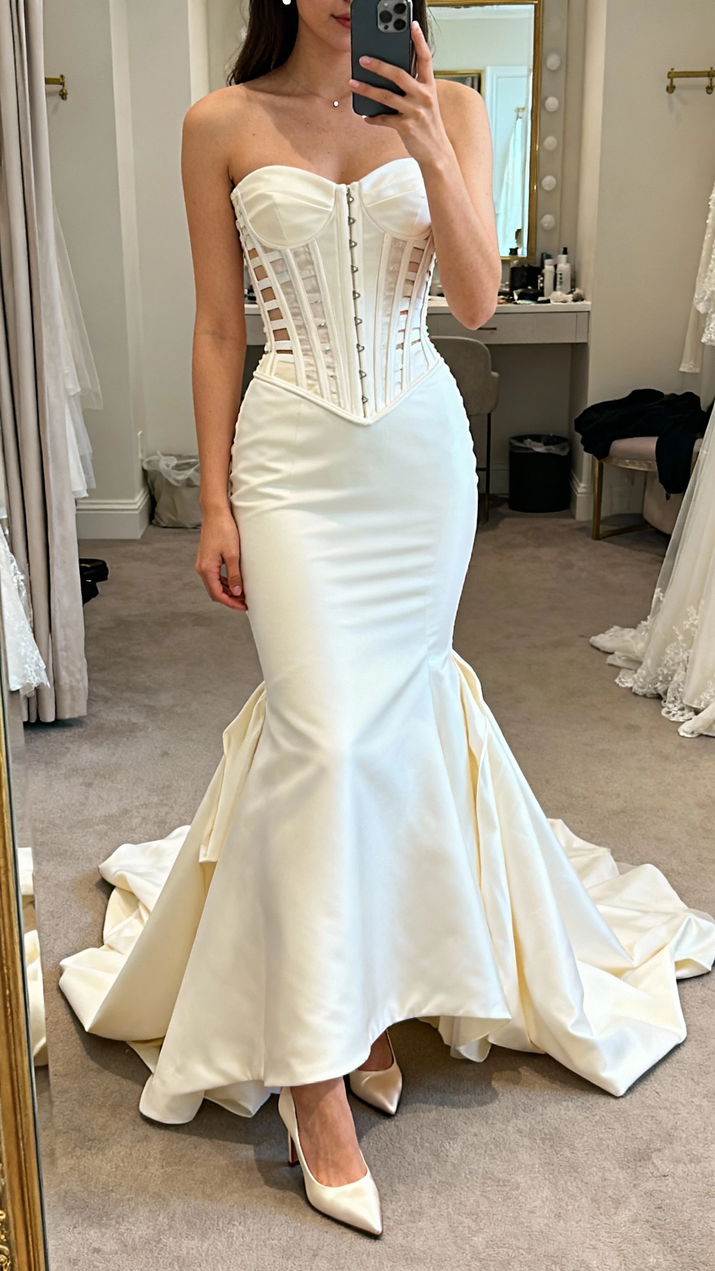 iPhone mirror selfie of a woman wearing a corseted mermaid wedding dress with visible boning and a detachable overskirt for a two-look effect, satin pumps and simple studs, face not visible, bridal suite mirror, soft indoor lighting, natural iPhone quality