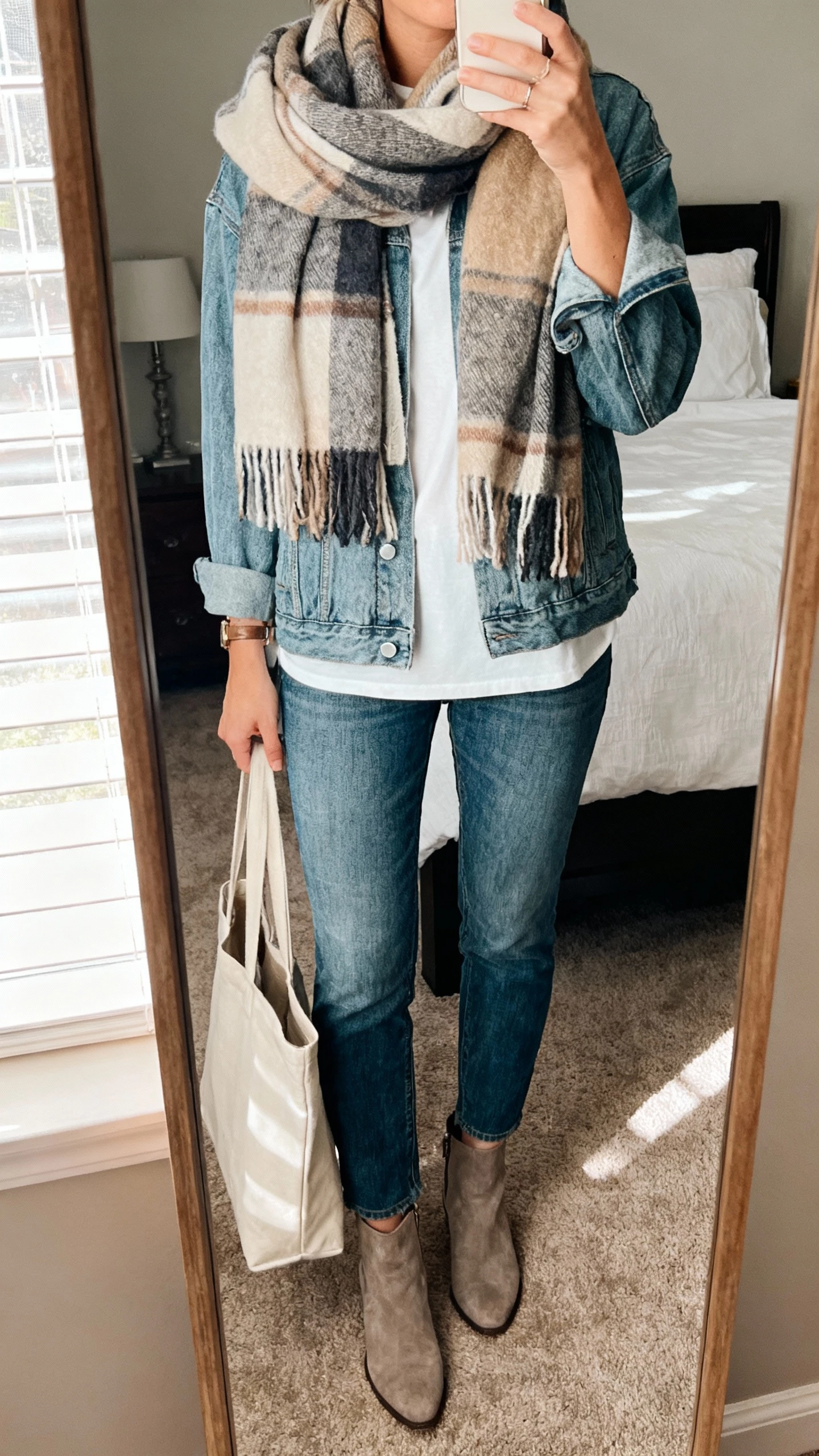 iPhone mirror selfie of a woman wearing a cozy blanket scarf over a relaxed denim jacket, straight-leg jeans, white tee, ankle boots, simple tote, face not visible, cropped at neck, bedroom mirror with morning window light, natural iPhone photo quality