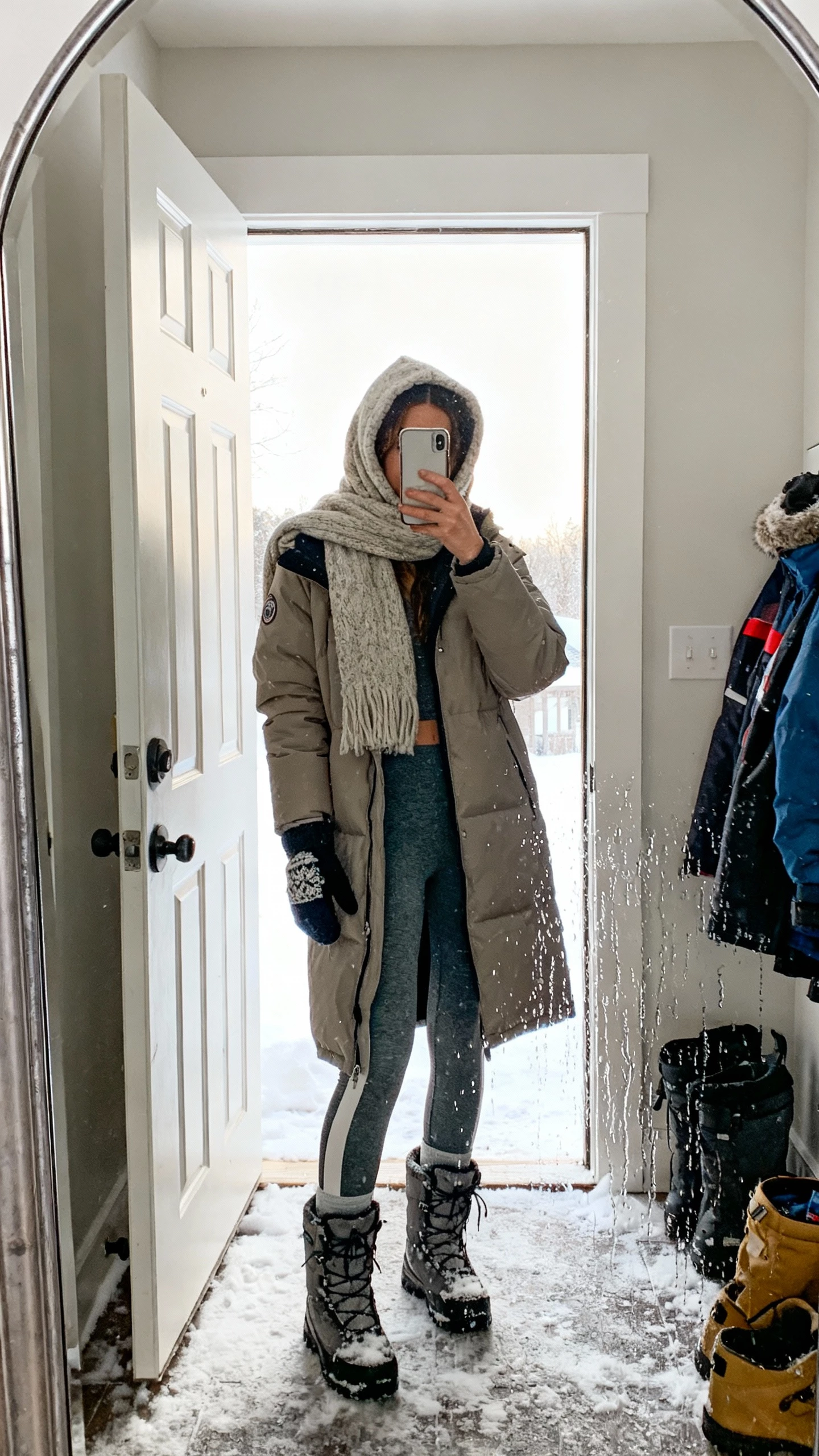 iPhone mirror selfie of a woman wearing a cozy hooded scarf pulled up over the head with a longline parka, thermal leggings, snow boots, and mittens, face not visible, entryway mirror with wet snow gear, bright daylight from open door, natural iPhone photo quality.