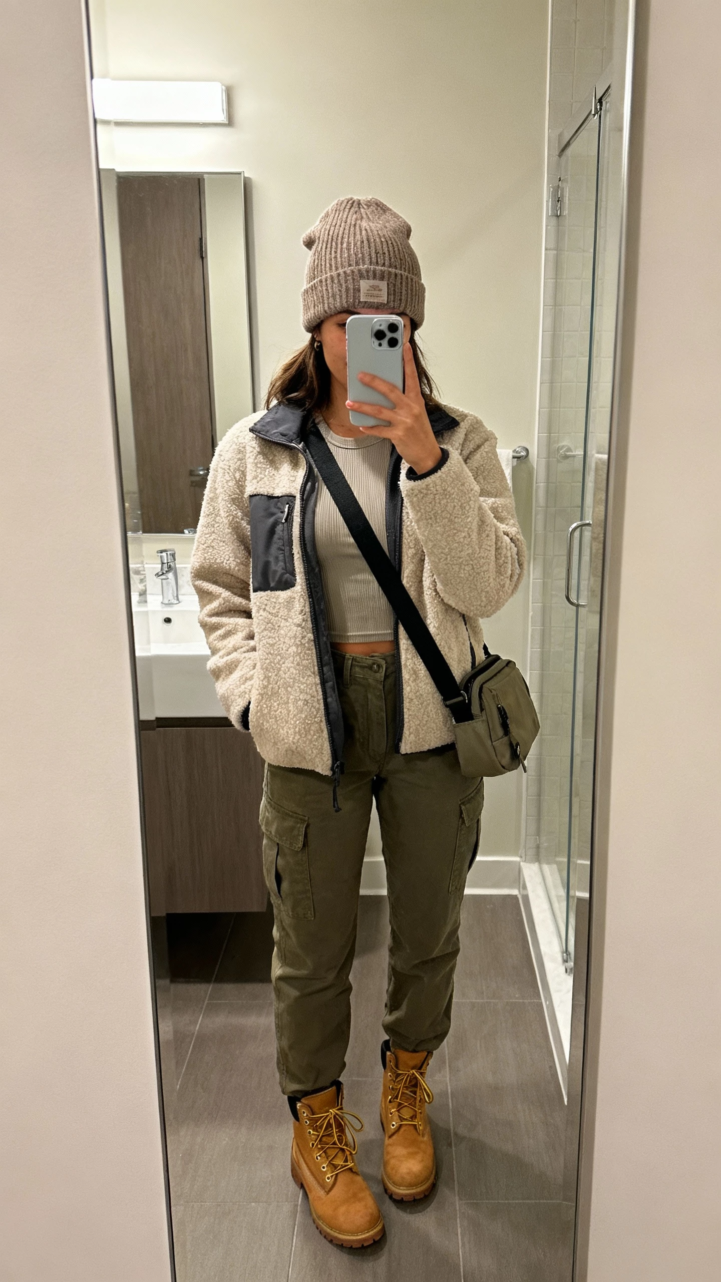 iPhone mirror selfie of a woman wearing a cozy shacket over a thermal top with cargo pants and lace-up boots, knit beanie, sling bag, face not visible, bathroom mirror with soft indoor lighting, natural iPhone photo quality.