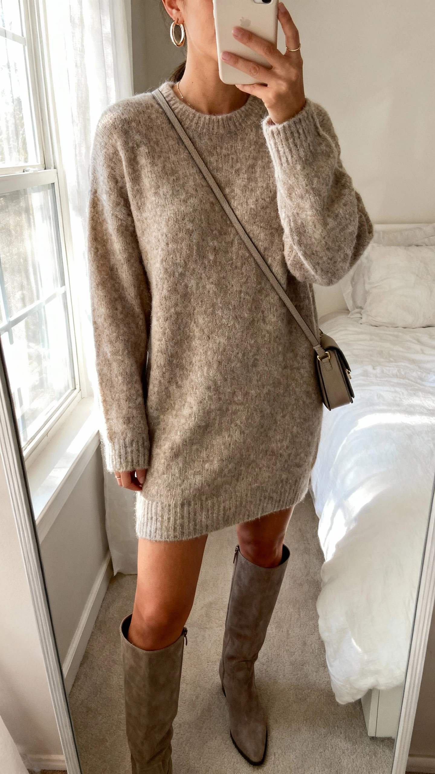 iPhone mirror selfie of a woman wearing a cozy sweater dress with knee-high boots, simple hoop earrings and a small crossbody bag, face not visible, cropped at neck, bedroom mirror with soft window light, natural iPhone photo quality.