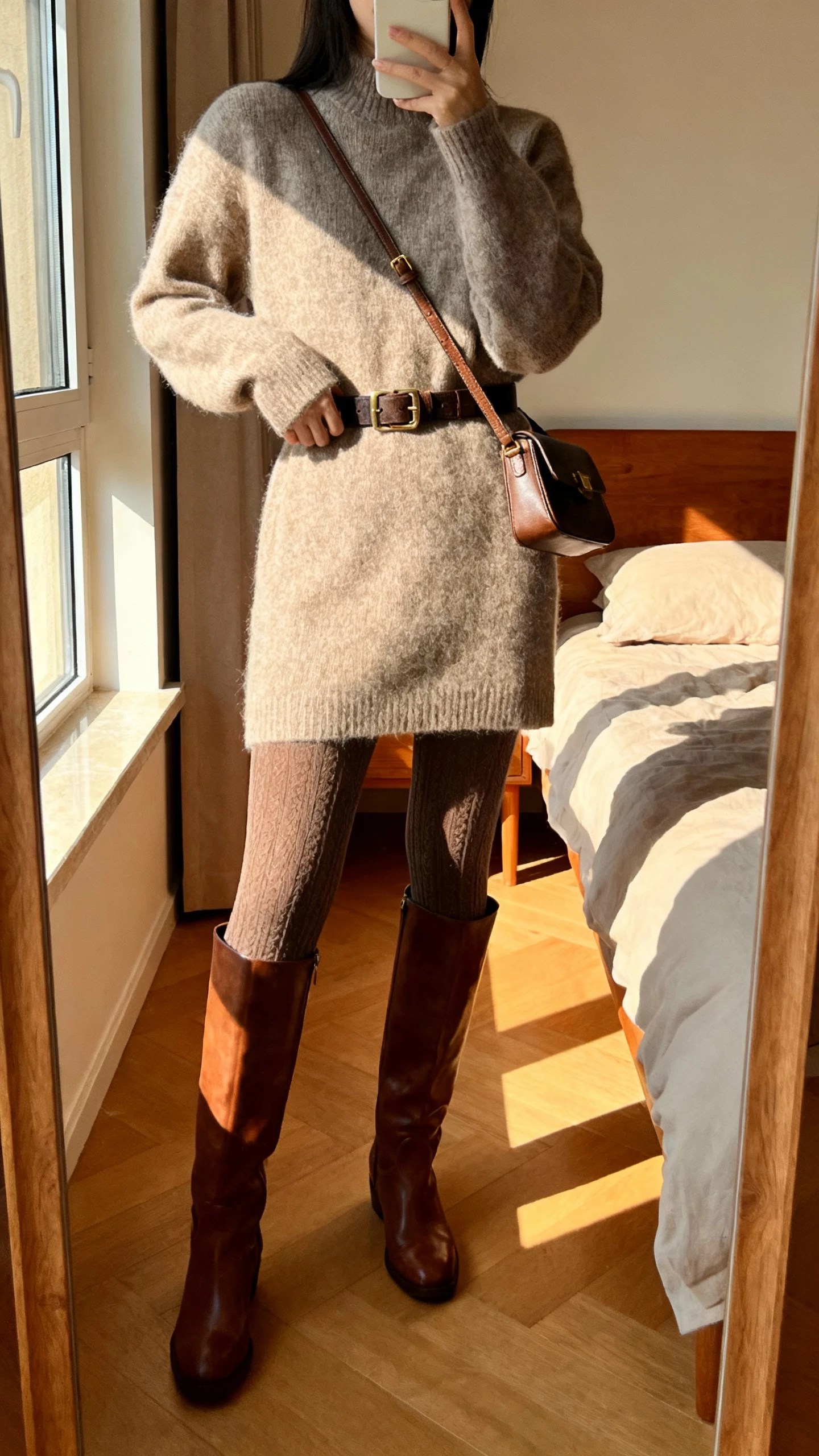 iPhone mirror selfie of a woman wearing a cozy sweater dress with knee-high leather boots, belt at waist, textured tights, small shoulder bag, face not visible, bedroom mirror, warm afternoon window light, natural iPhone photo quality