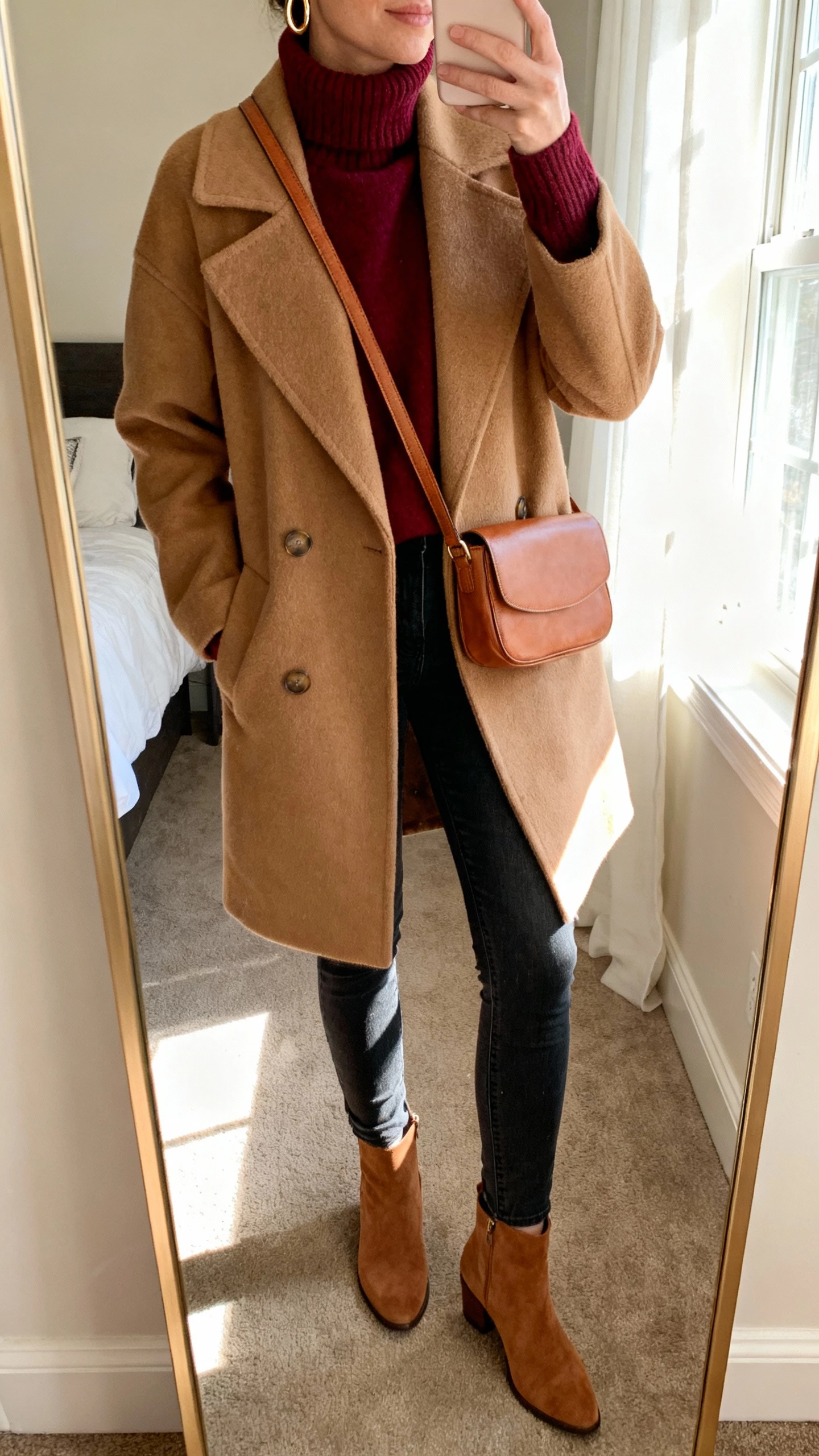 iPhone mirror selfie of a woman wearing a cranberry turtleneck sweater, camel wool coat, slim dark jeans, camel ankle boots, camel crossbody bag, simple gold hoops, face not visible, cropped at neck, bedroom mirror with morning window light, natural iPhone photo quality