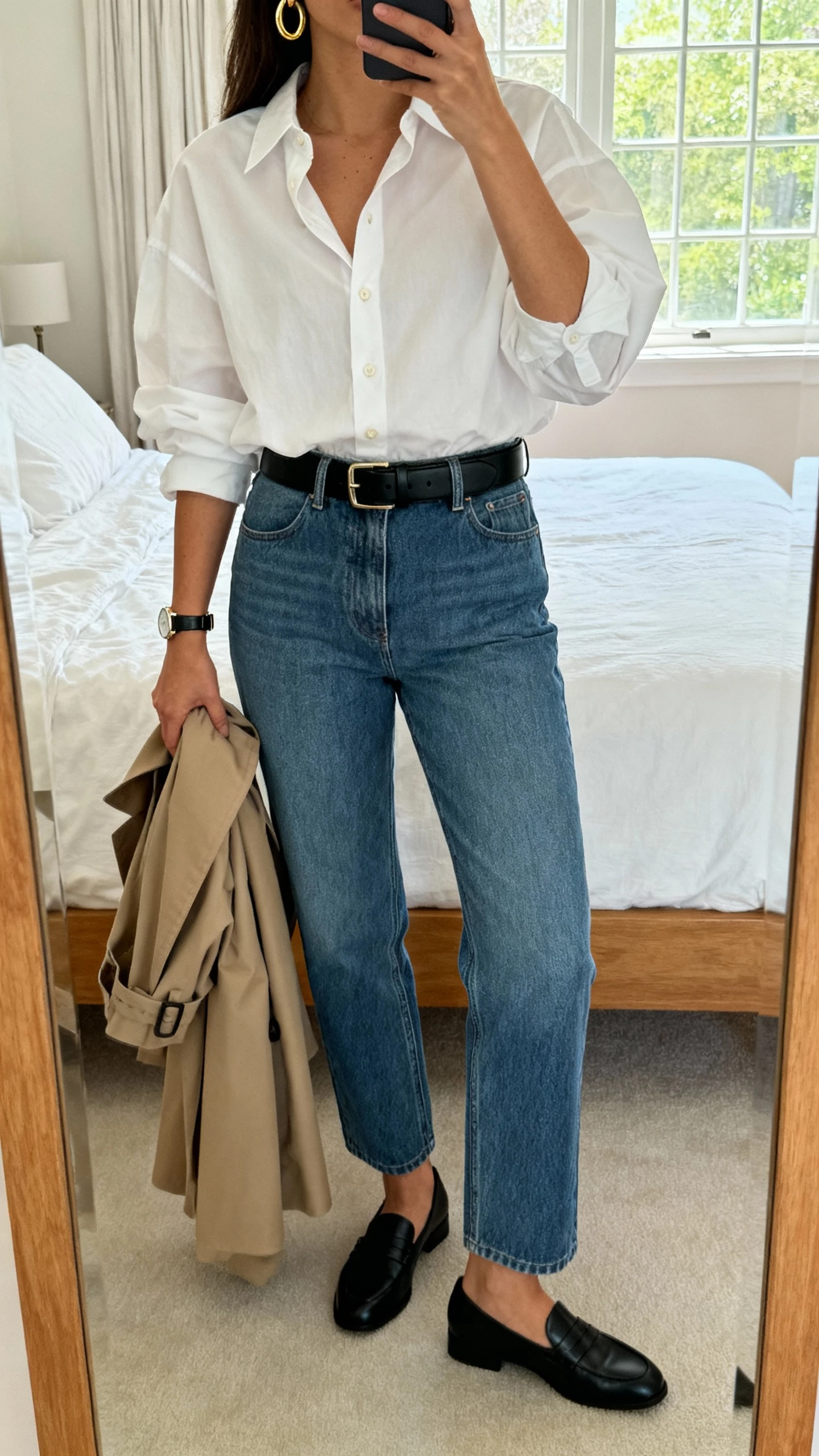 iPhone mirror selfie of a woman wearing a crisp white button-down (slightly oversized), mid-rise straight-leg blue jeans, black loafers, structured black belt, gold hoop earrings, minimal watch, optional trench draped over arm; bedroom mirror, face not visible, natural window light, natural iPhone photo quality.
