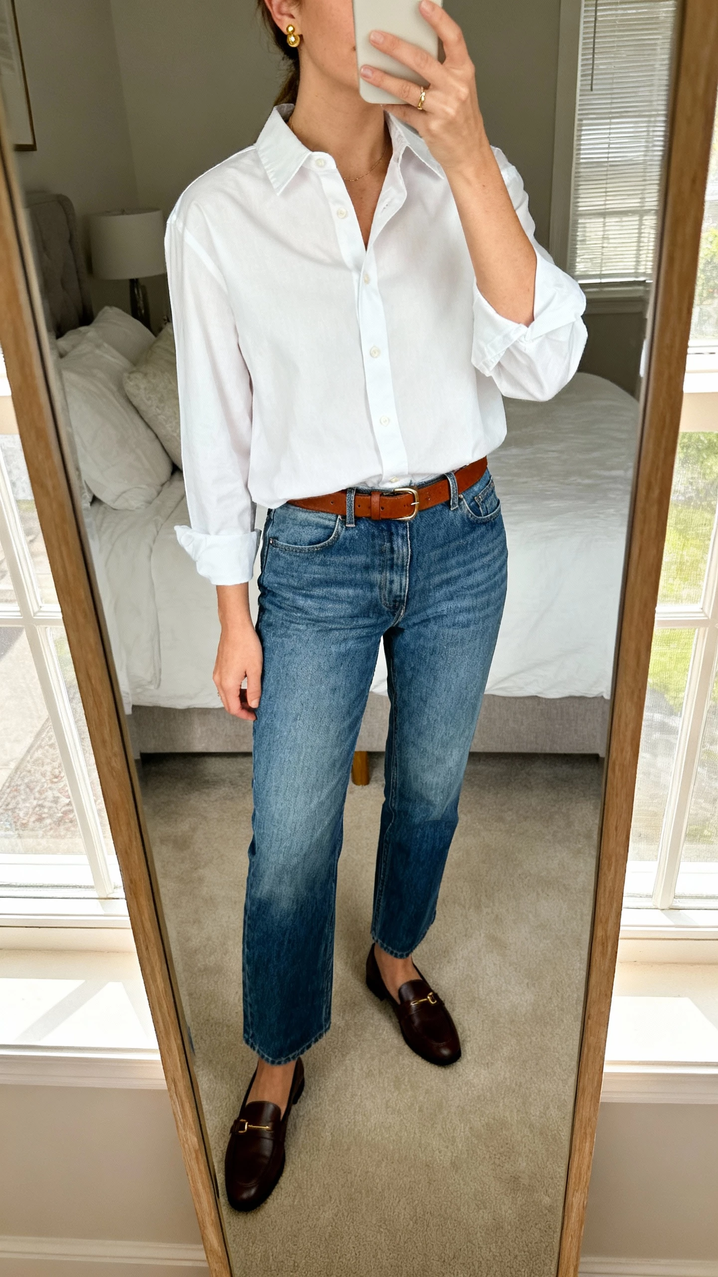 iPhone mirror selfie of a woman wearing a crisp white button-up shirt tucked into mid-wash blue straight jeans, brown belt, loafers, delicate gold studs, face not visible, cropped at neck, bedroom mirror with morning window light, natural iPhone photo quality.