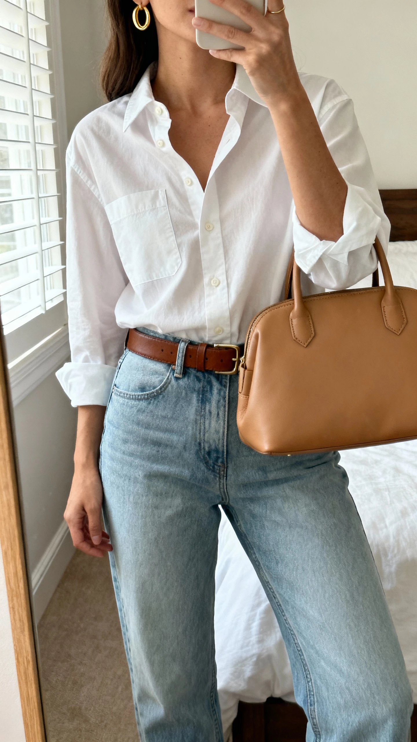 iPhone mirror selfie of a woman wearing a crisp white button-down tucked into light blue jeans, brown leather belt, simple gold hoops and a structured tan tote, face not visible, cropped at neck, bedroom mirror with morning window light, natural iPhone photo quality