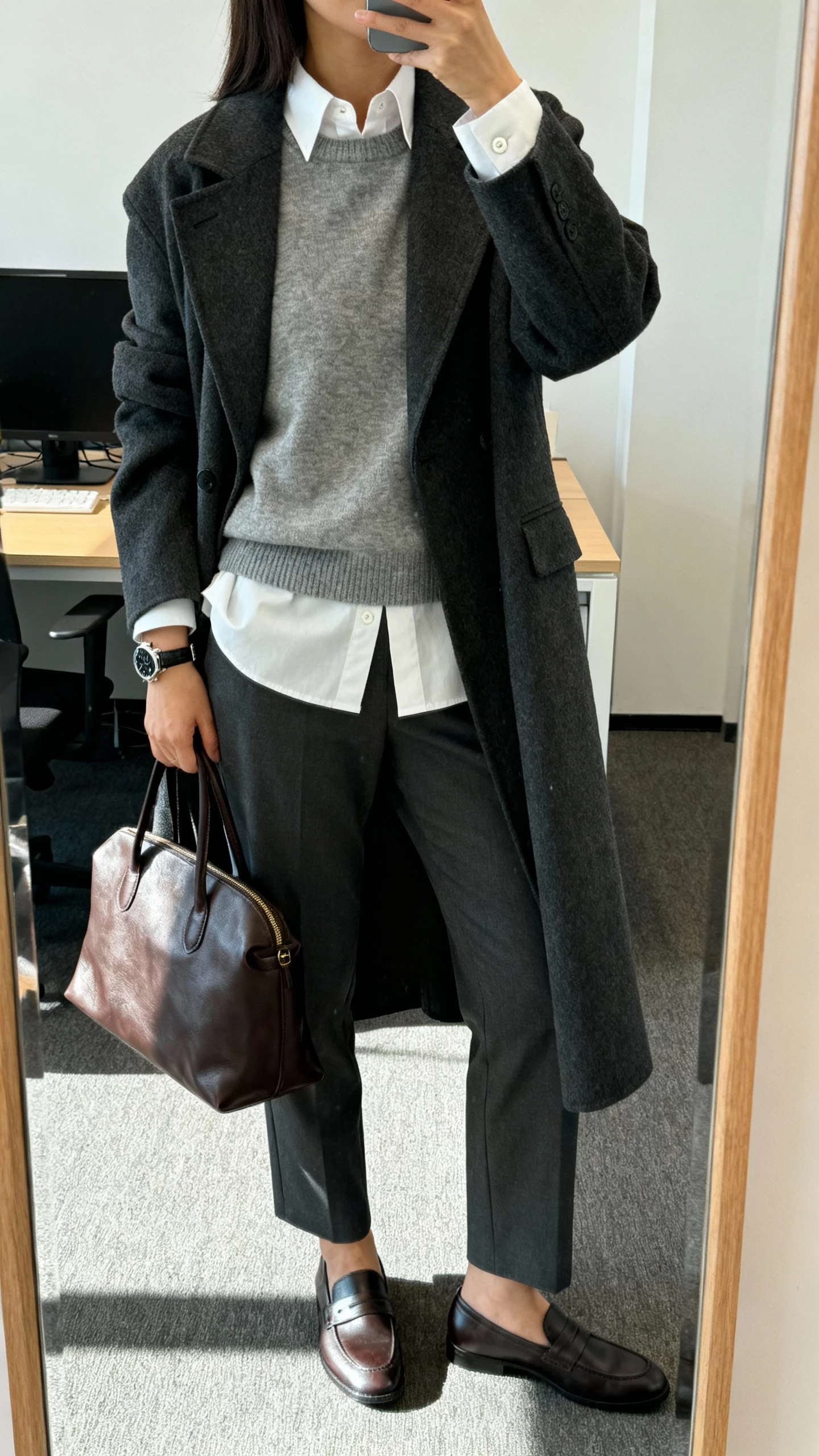 iPhone mirror selfie of a woman wearing a crisp white button-down under a heather grey crewneck sweater with a long menswear coat on top, straight-leg trousers, loafers, watch and leather tote, face not visible, cropped at neck, office mirror, morning light, casual iPhone quality.