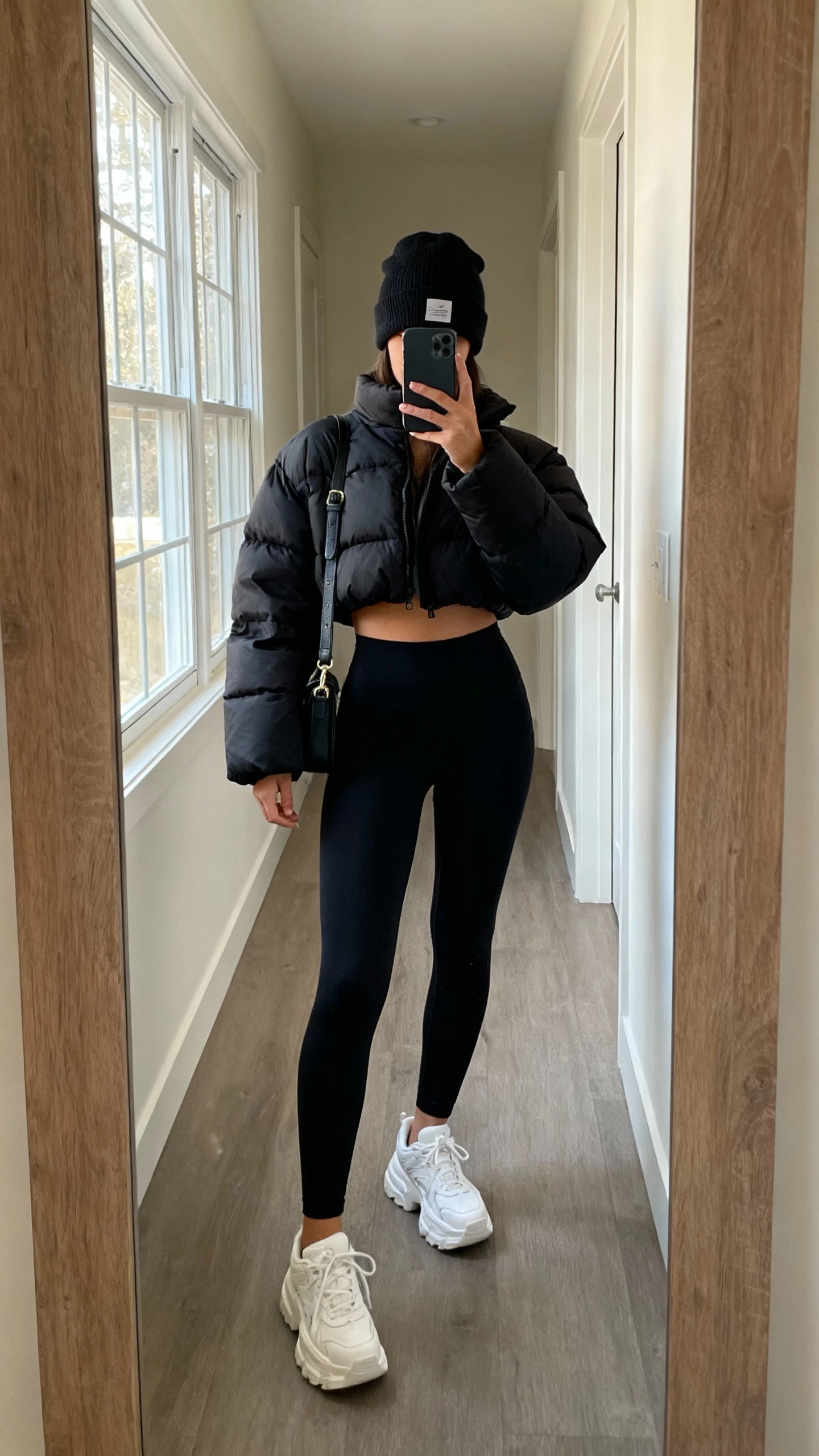 iPhone mirror selfie of a woman wearing a cropped black puffer jacket, high-waisted black leggings, chunky white sneakers, beanie, crossbody bag, face not visible, cropped at neck, hallway mirror, morning window light, natural iPhone photo quality