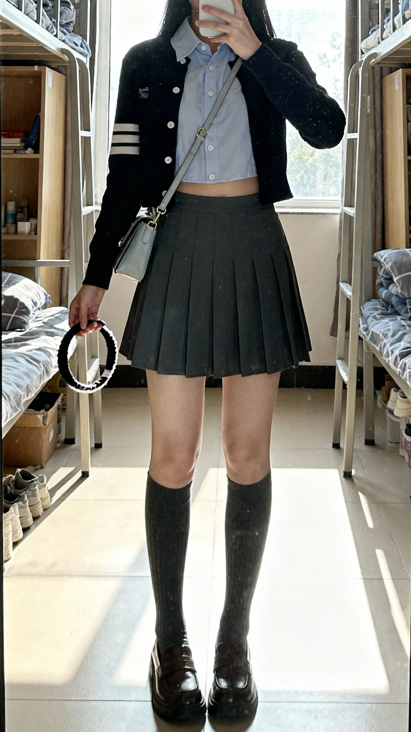 iPhone mirror selfie of a woman wearing a cropped cardigan layered over a crisp button-down with a pleated mini skirt, knee-high socks, loafers, headband in hand, small shoulder bag, face not visible, dorm-style bedroom setting, morning window light, iPhone photo quality.
