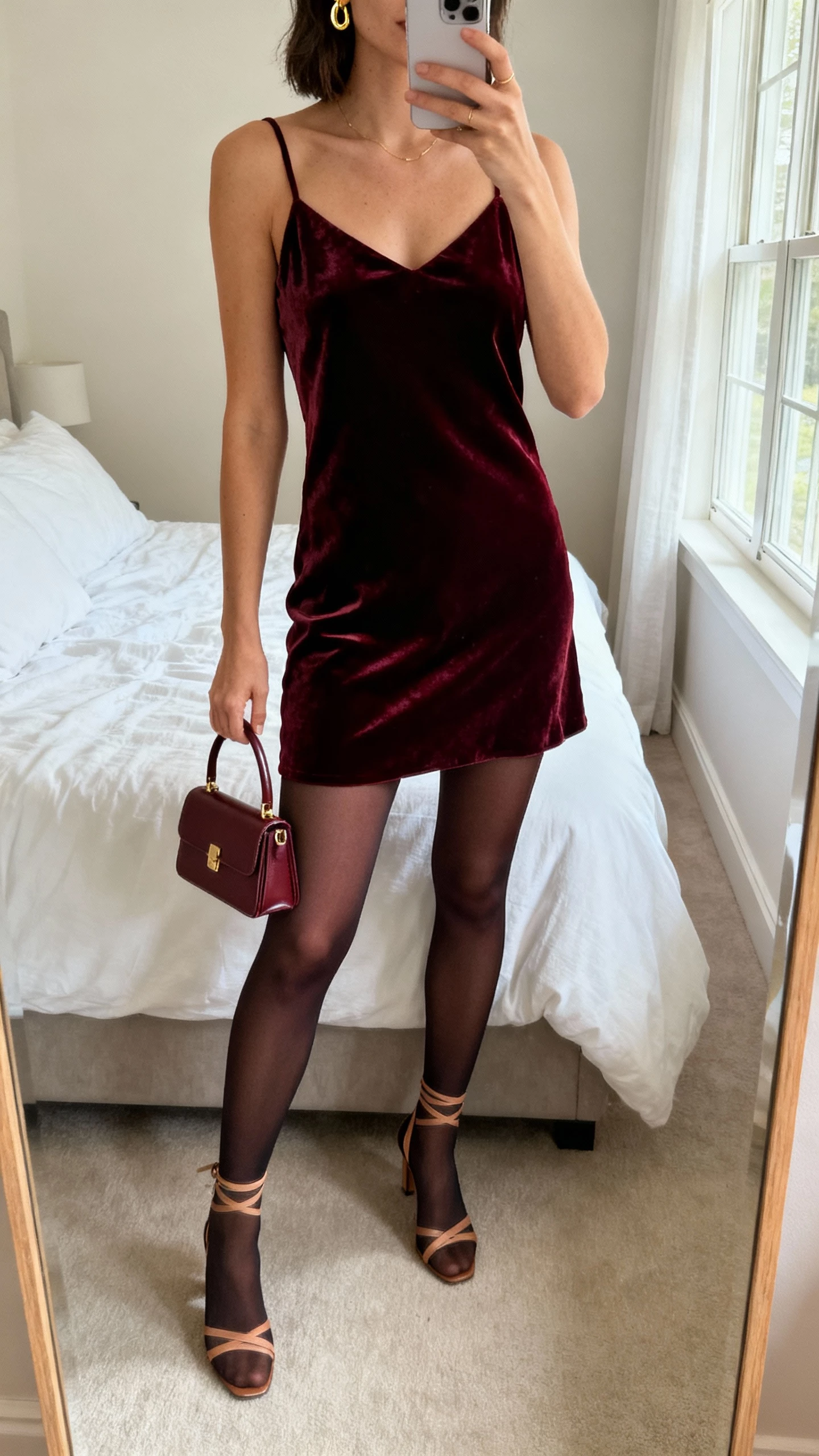 iPhone mirror selfie of a woman wearing a deep burgundy velvet slip dress layered over opaque power tights, strappy heels, delicate gold earrings, mini top-handle bag, face not visible, cropped at neck, bedroom mirror with soft window light, natural iPhone photo quality.