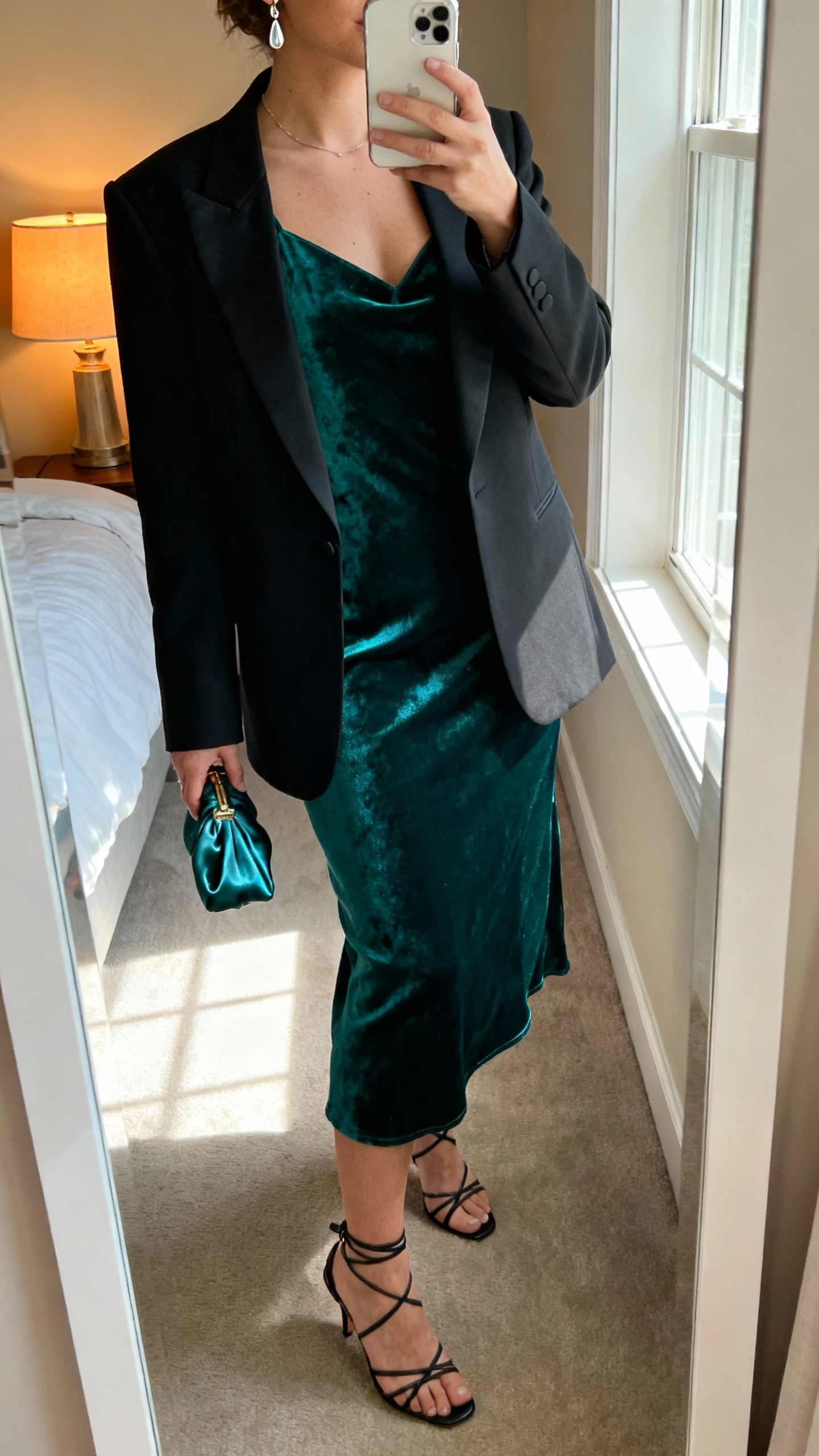 iPhone mirror selfie of a woman wearing a deep emerald velvet slip dress with a black tuxedo blazer, strappy heels, delicate drop earrings, and a small satin clutch, face not visible, cropped at neck, bedroom mirror with warm lamp and window light, natural iPhone photo quality.