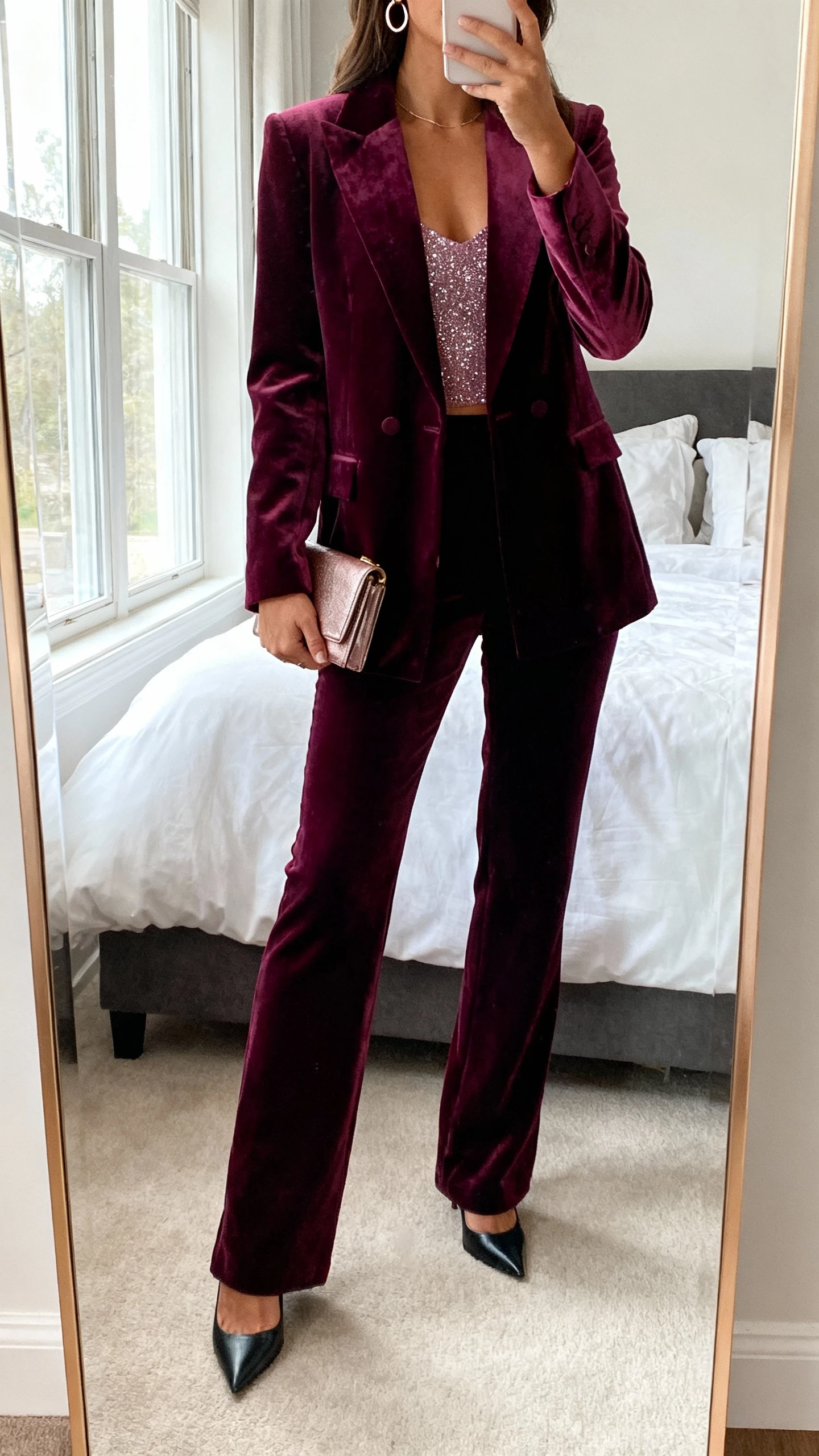 iPhone mirror selfie of a woman wearing a deep jewel-tone velvet power suit with a subtle sparkle cami, pointed heels, delicate hoop earrings, and a mini clutch, face not visible, cropped at neck, bedroom full-length mirror, soft window light, natural iPhone photo quality.
