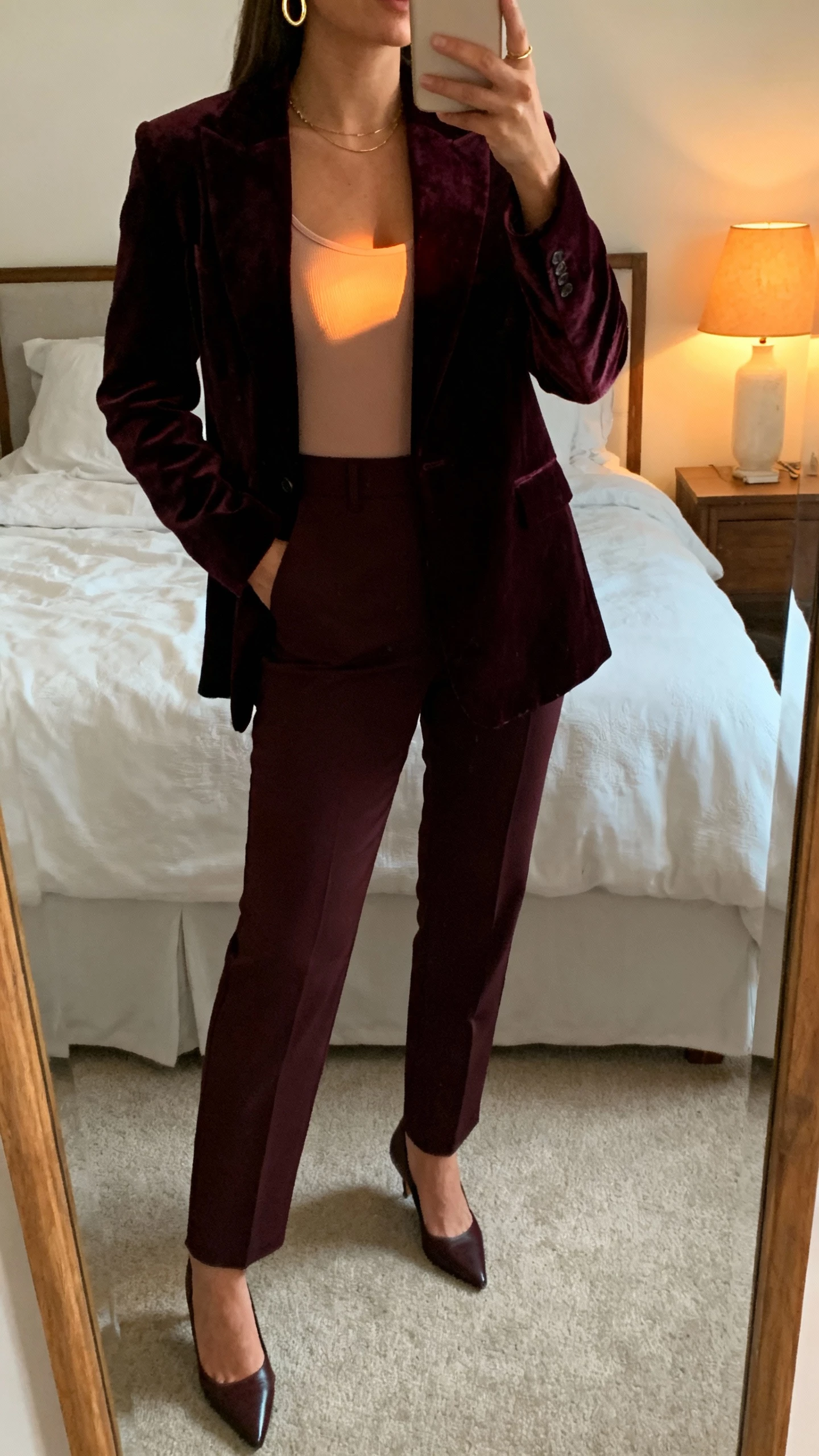 iPhone mirror selfie of a woman wearing a deep velvet suit (blazer and tailored trousers) with a soft-glow cami, delicate gold hoops, pointed heels, face not visible, cropped at neck, bedroom mirror with warm lamp light, natural iPhone photo quality.
