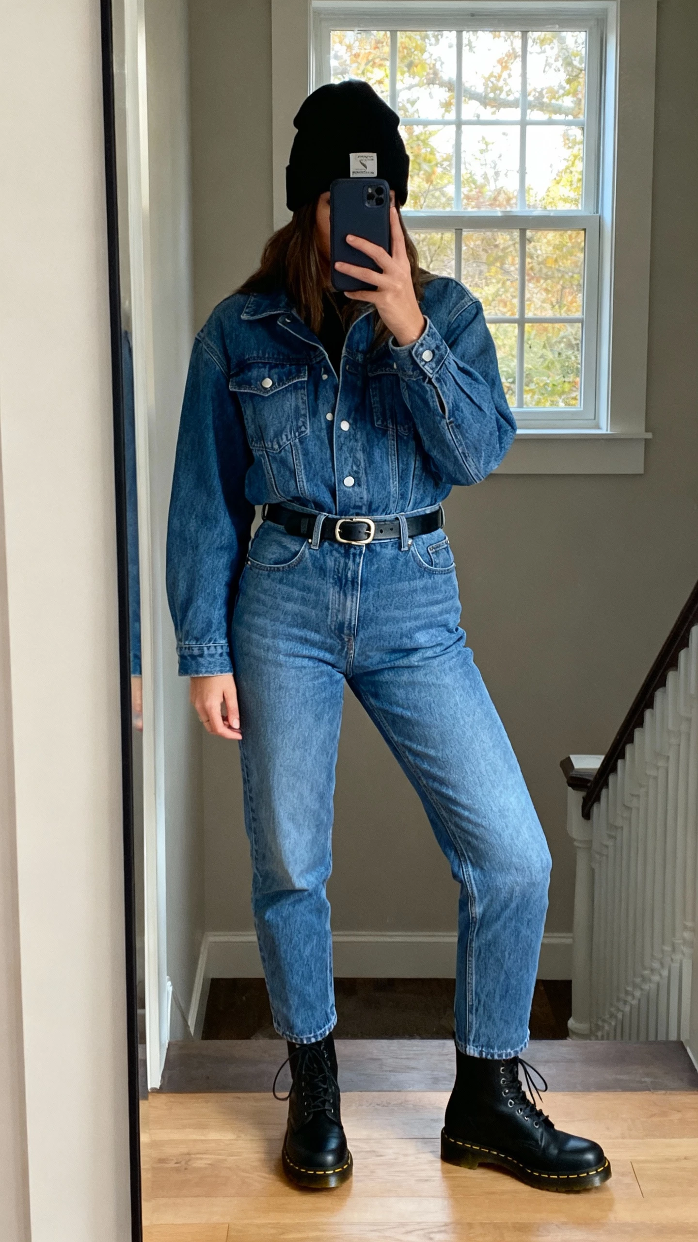 iPhone mirror selfie of a woman wearing a denim jacket with matching high-waisted jeans, black Doc Martens, a black beanie, simple belt, face not visible, entryway mirror by a window, natural afternoon light, natural iPhone photo quality.
