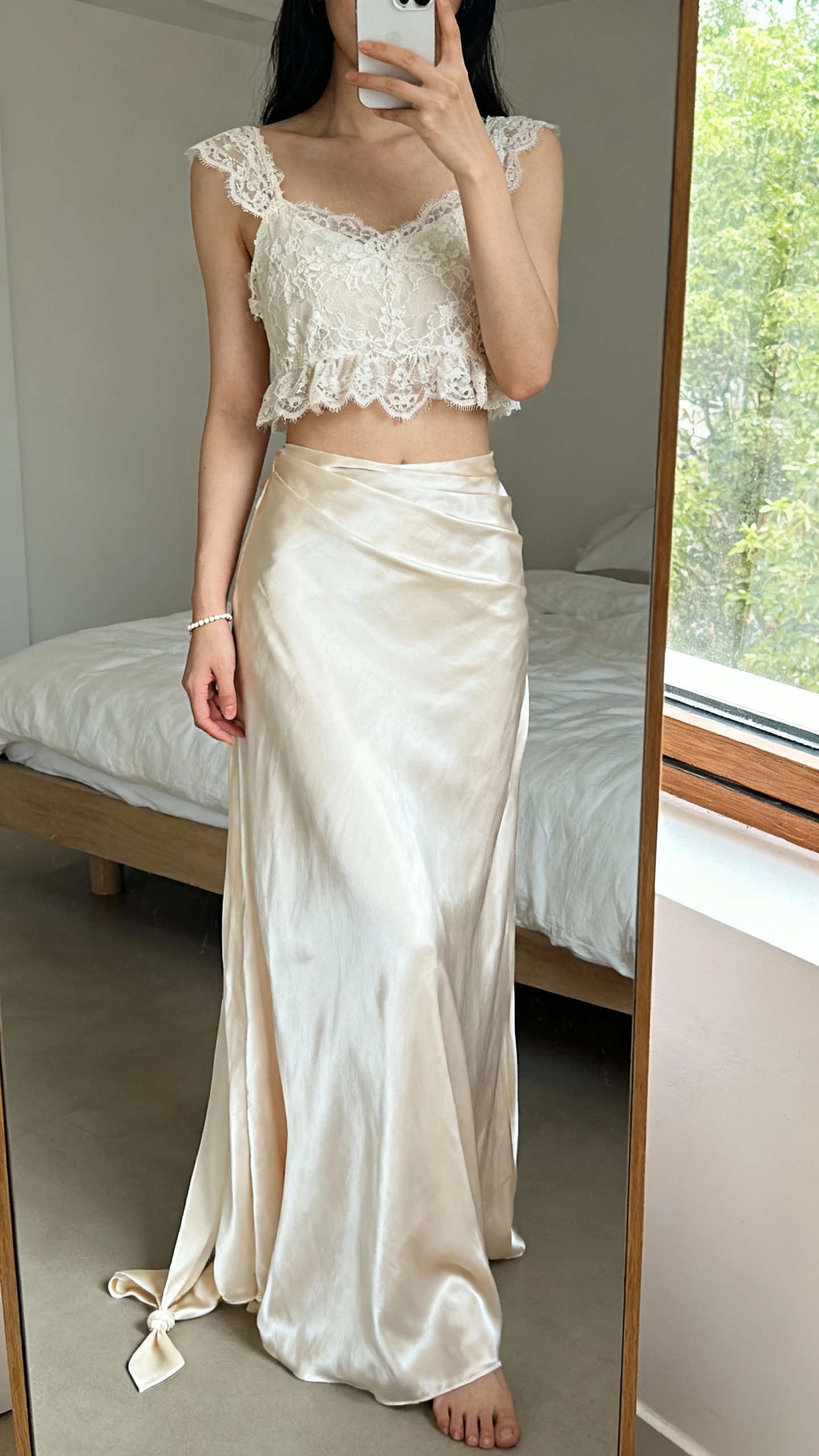iPhone mirror selfie of a woman wearing a dreamy two-piece boho wedding set: cropped lace top and flowing silk skirt with a small train, delicate bracelet, face not visible, minimalist bedroom mirror, morning window light, natural iPhone photo quality