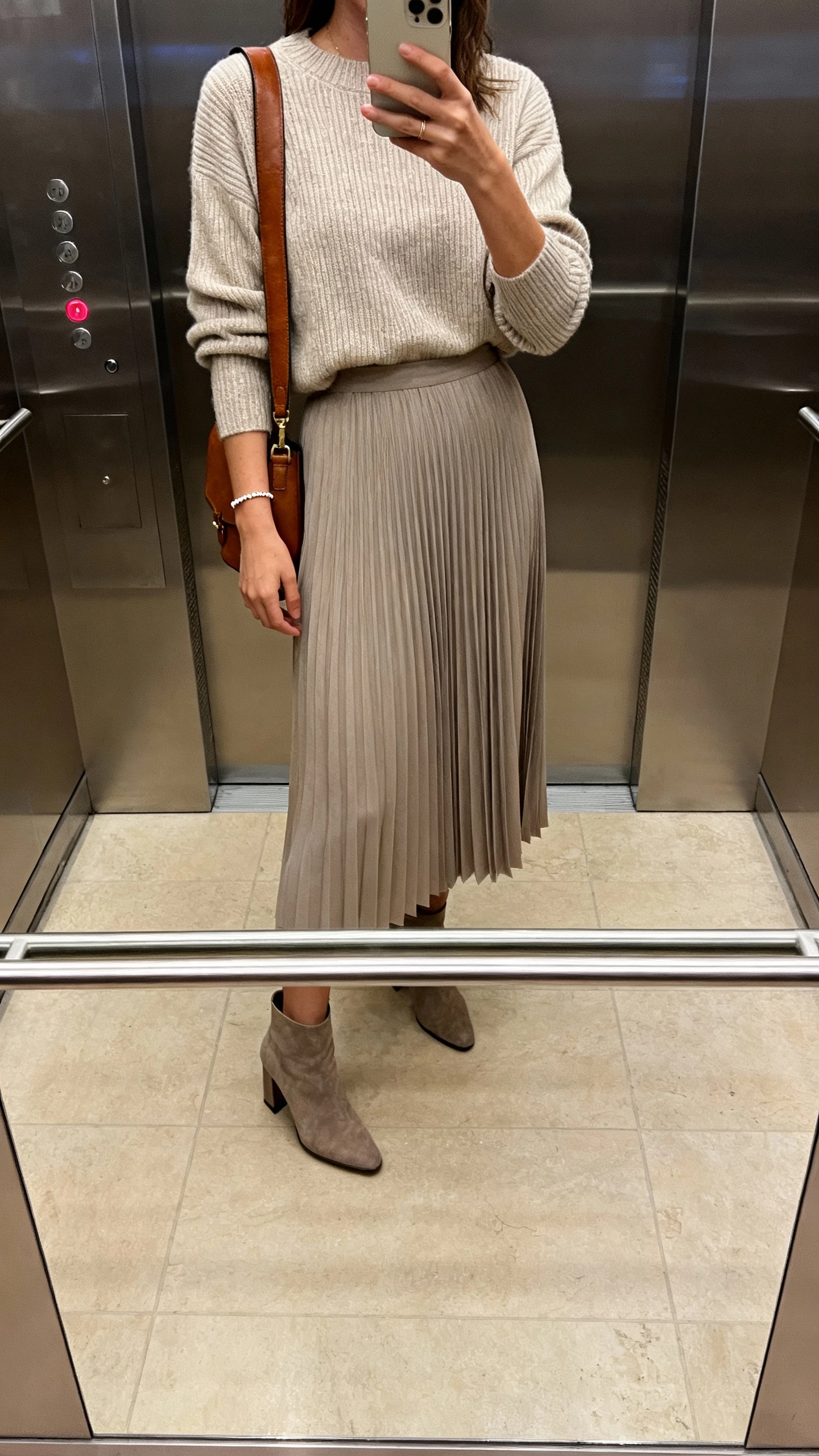 iPhone mirror selfie of a woman wearing a fine-gauge knit sweater tucked into a midi pleated skirt, heeled ankle boots, delicate bracelet, leather shoulder bag, face not visible, elevator mirror after work, soft evening light, natural iPhone photo.