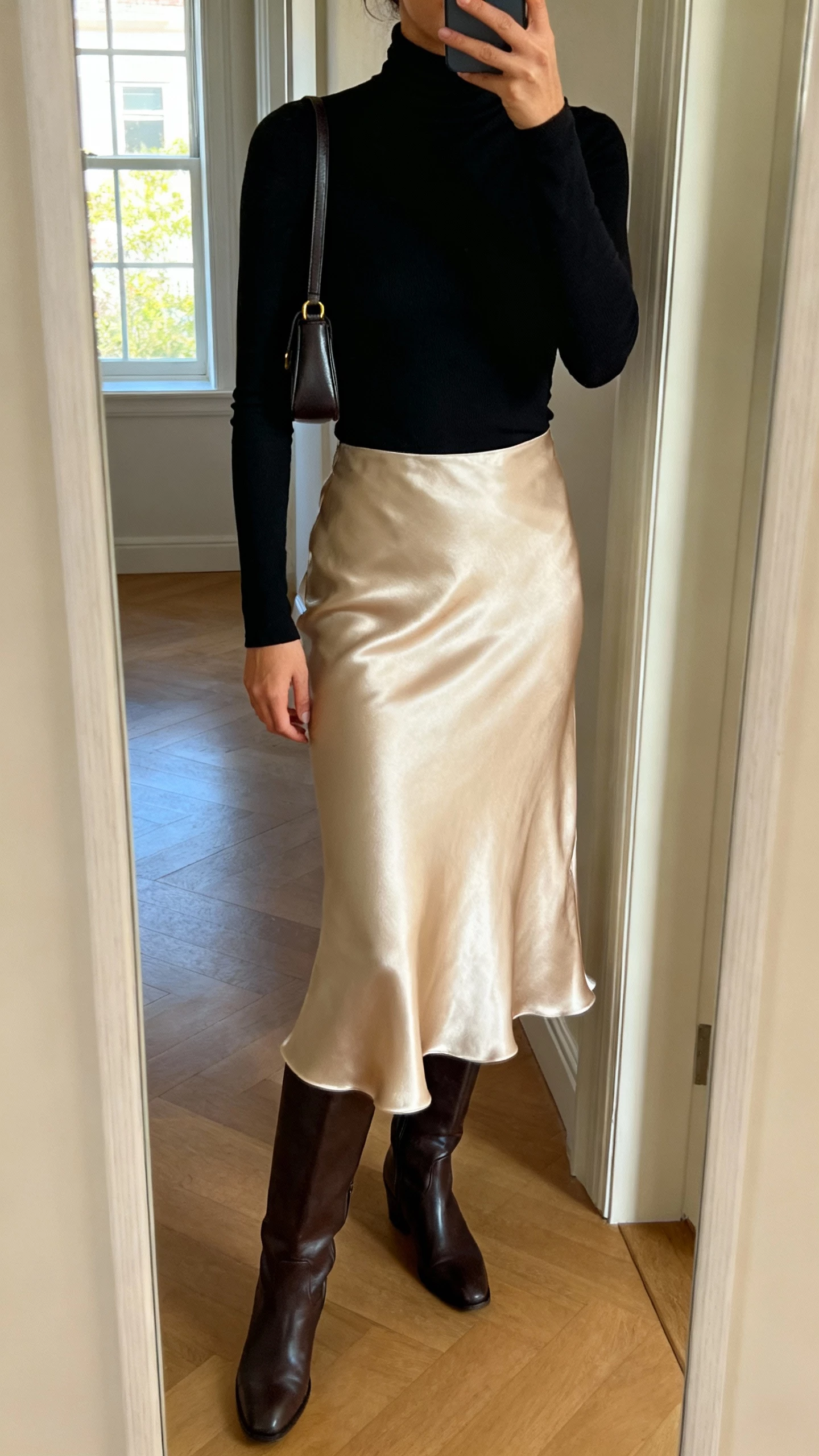 iPhone mirror selfie of a woman wearing a fitted black turtleneck, satin slip skirt in champagne, knee-high leather boots, small shoulder bag, face not visible, cropped at shoulders, entryway mirror, daylight from window, natural iPhone quality