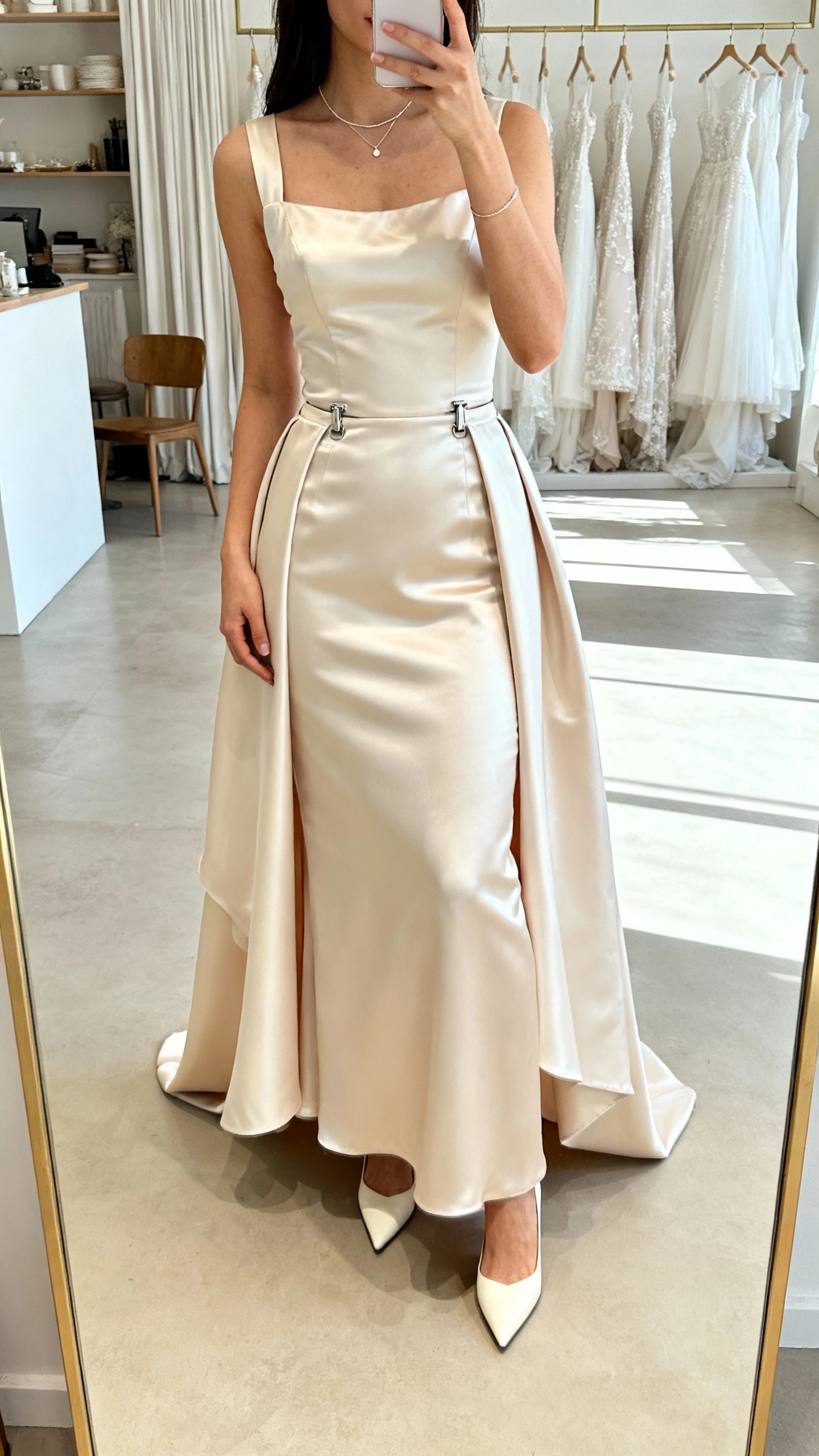 iPhone mirror selfie of a woman wearing a fitted satin gown with a detachable overskirt clipped at the waist for two-look styling, minimalist jewelry, and pointed heels, face not visible, bridal studio mirror, bright indoor lighting, natural iPhone photo
