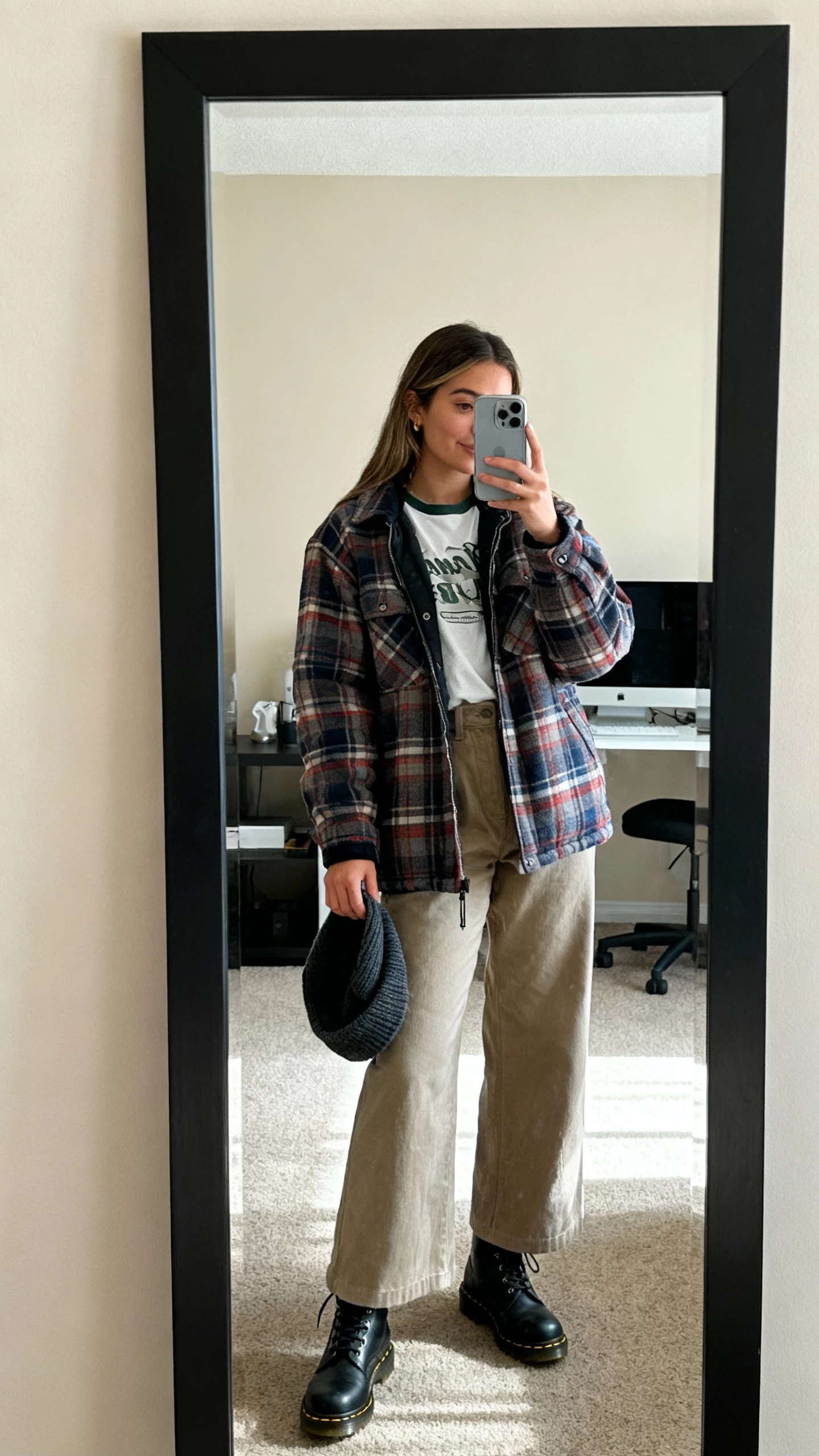 iPhone mirror selfie of a woman wearing a flannel shacket over a tee, wide-leg pants, Doc Martens, beanie in hand, face not visible, studio apartment mirror, soft daylight, casual iPhone photo.