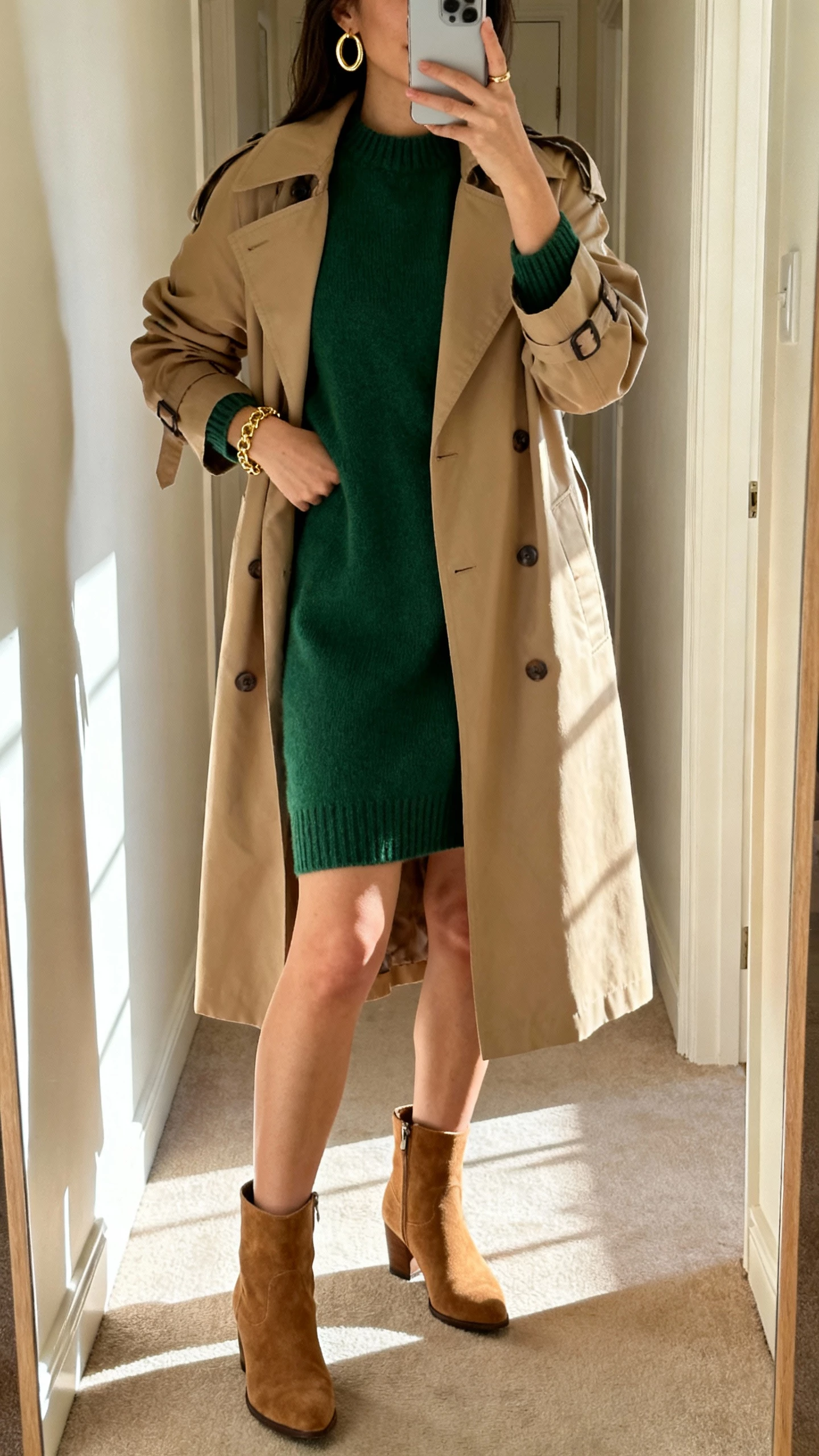 iPhone mirror selfie of a woman wearing a forest green sweater dress, sand trench coat, gold hoop earrings and bracelet, tan suede ankle boots, face not visible, hallway mirror with morning light, natural iPhone photo quality