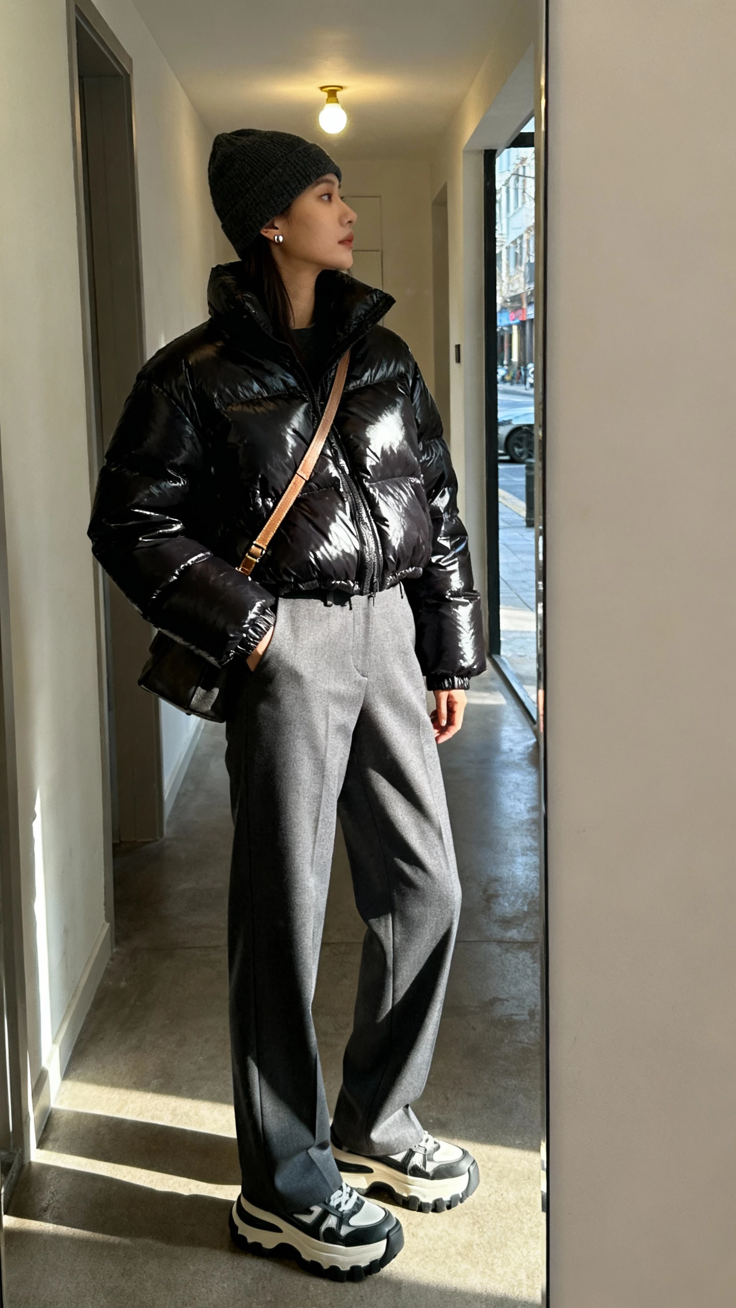 iPhone mirror selfie of a woman wearing a glossy black puffer jacket over tailored gray trousers, chunky-soled sneakers, crossbody bag, beanie, and minimalist studs, face not visible, cropped at neck in a hallway mirror with street light spilling in, natural iPhone photo quality.