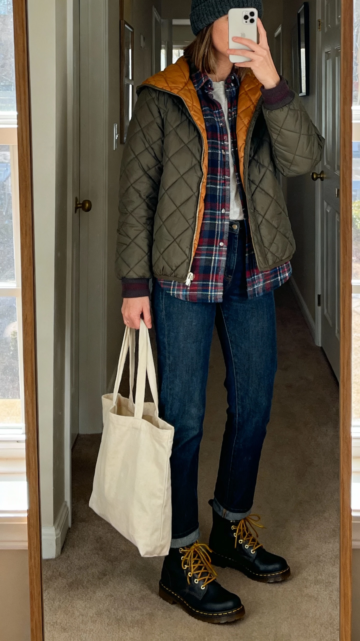 iPhone mirror selfie of a woman wearing a layered flannel shirt under a quilted liner jacket, straight-leg jeans, lace-up boots, beanie, and canvas tote, face not visible, cropped at shoulders, hallway mirror in an apartment with morning window light, natural iPhone photo quality.