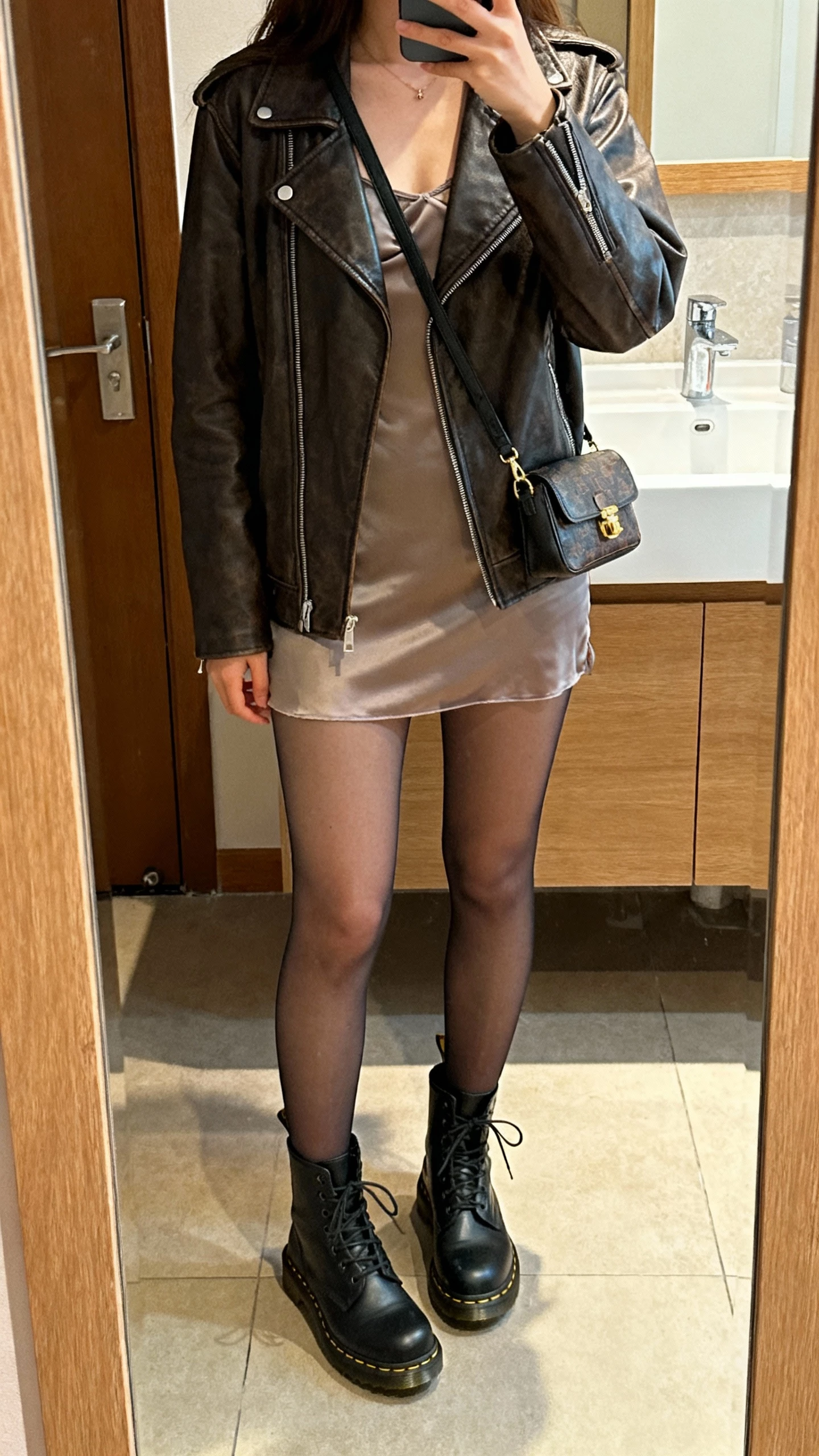 iPhone mirror selfie of a woman wearing a leather jacket over a slip dress with opaque tights and Doc Martens, small shoulder bag, face not visible, bathroom mirror, warm indoor lighting, natural iPhone photo quality.
