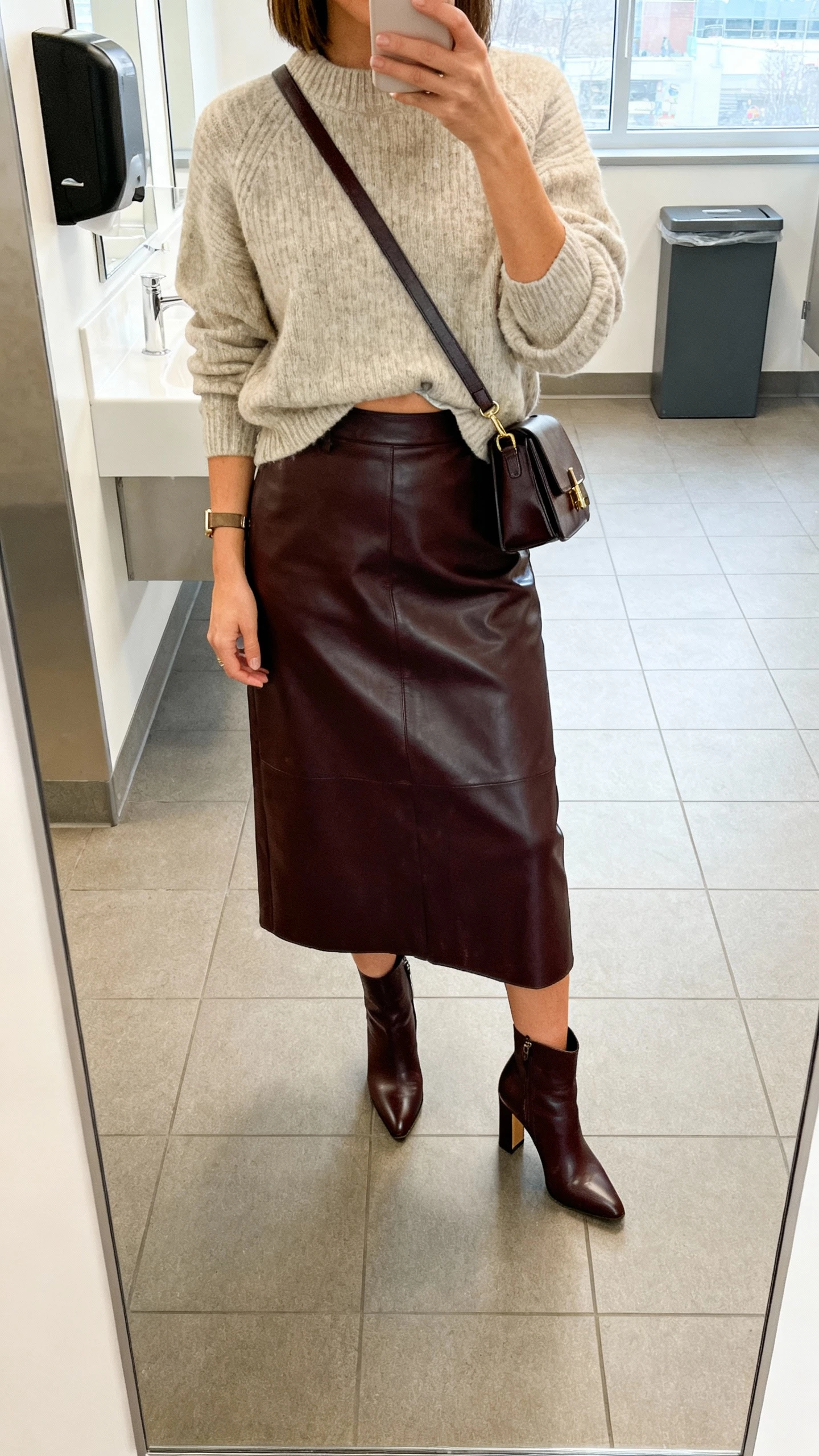 iPhone mirror selfie of a woman wearing a leather midi skirt with a soft knit sweater half-tucked, heeled ankle boots, and a structured crossbody, face not visible, office restroom mirror, natural overhead and window light, iPhone photo quality.
