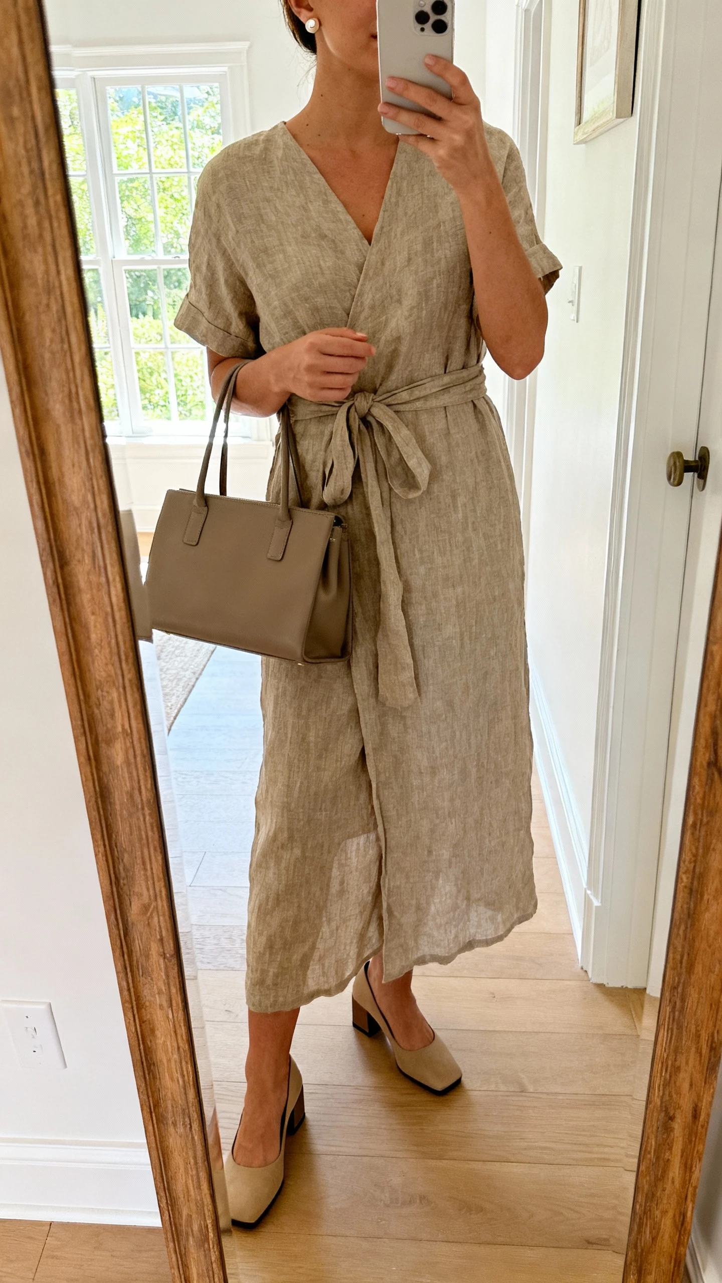 iPhone mirror selfie of a woman wearing a linen-blend wrap dress, low block heels, structured tote, simple stud earrings, face not visible, cropped at shoulders, entryway mirror, bright afternoon window light, natural iPhone photo.