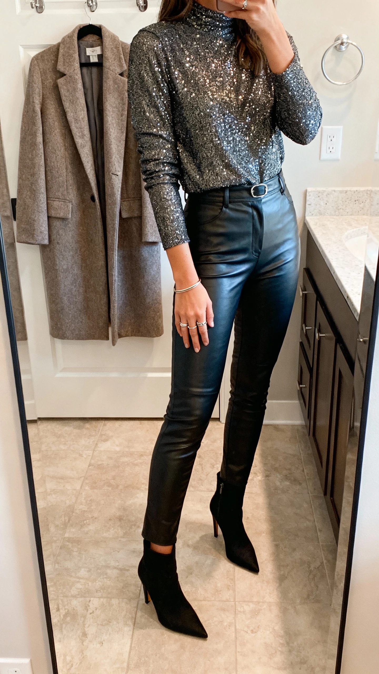 iPhone mirror selfie of a woman wearing a long-sleeve sequin top tucked into high-waisted faux-leather slim pants, pointed heeled booties, delicate stacked rings and a slim bangle, tailored wool-blend coat on hook behind, face not visible, bathroom mirror under soft indoor lighting, natural iPhone photo.