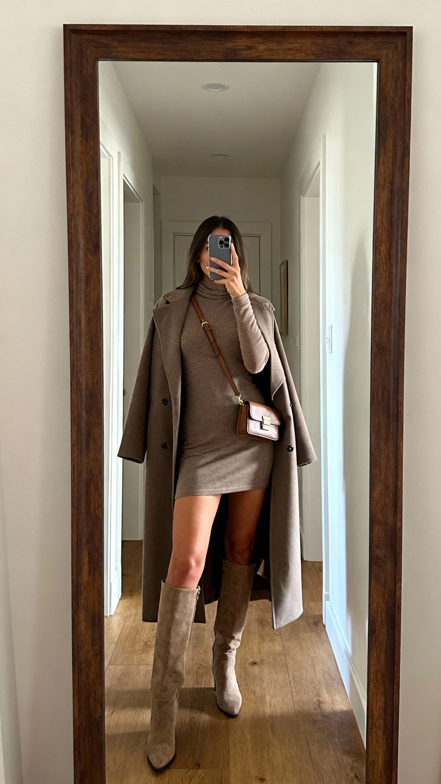 iPhone mirror selfie of a woman wearing a long-sleeve mini dress with tall knee-high boots and a structured coat draped like a dress, small crossbody bag, face not visible, entryway full-length mirror, natural iPhone photo quality
