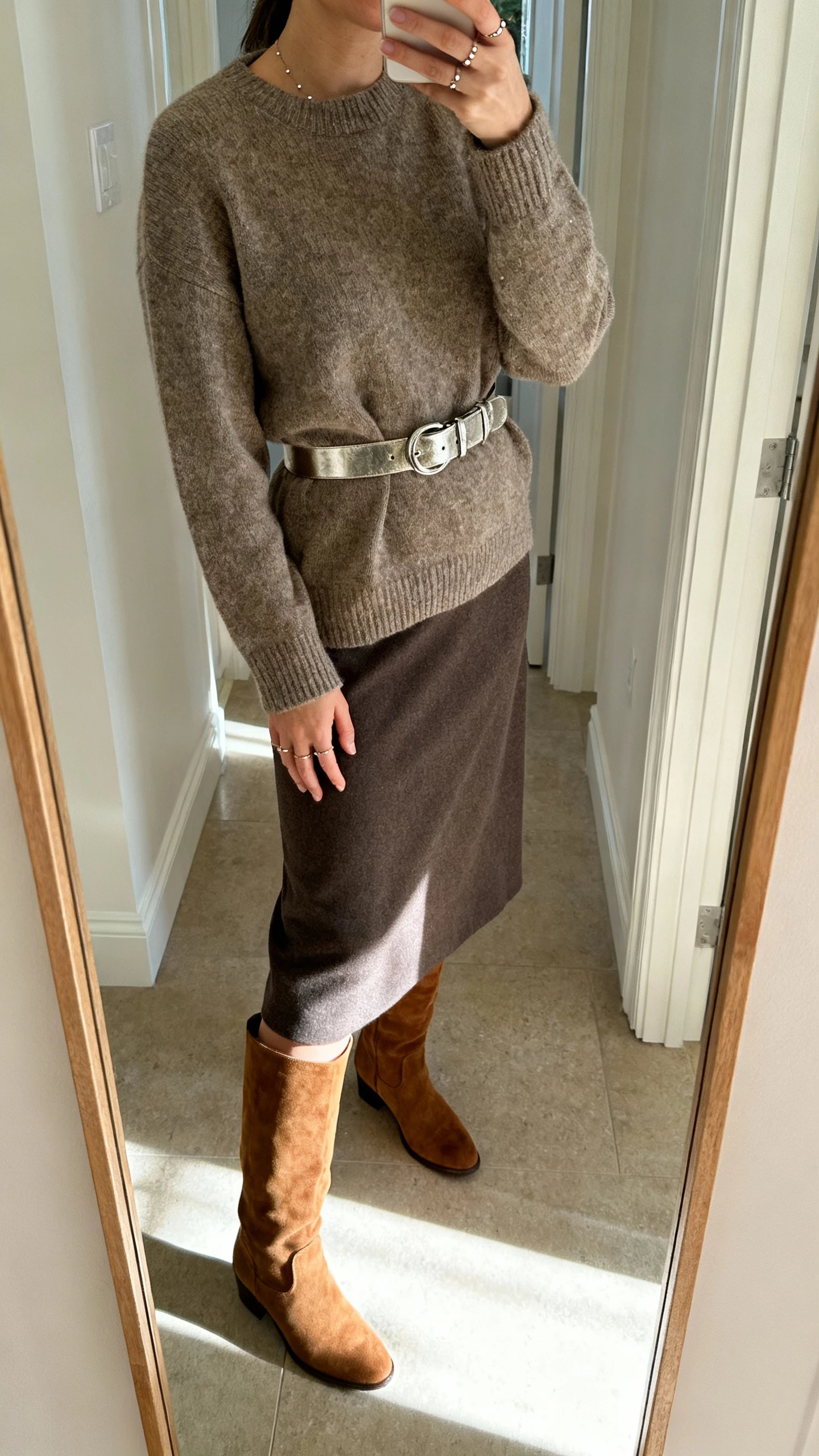 iPhone mirror selfie of a woman wearing a metallic belt cinching a tailored sweater and midi skirt, suede knee-high boots, delicate studs, face not visible, cropped at neck, clean entryway mirror with soft morning light, natural iPhone photo quality.