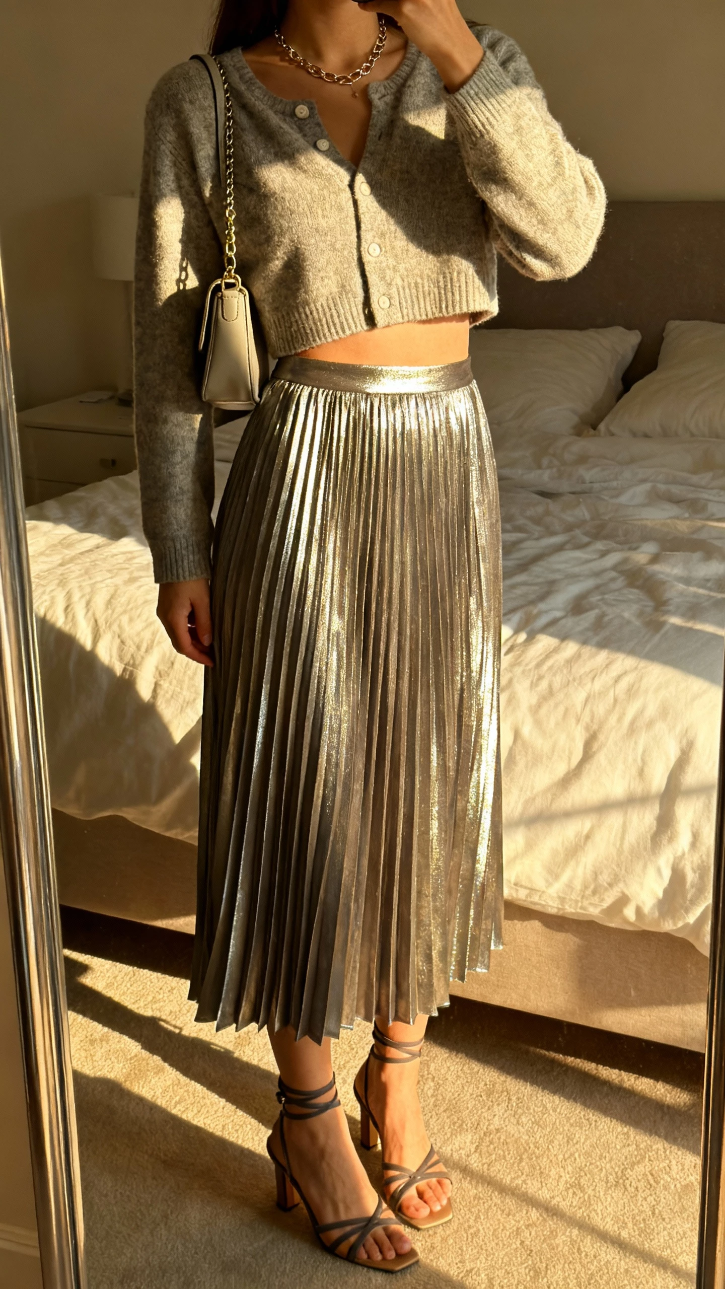 iPhone mirror selfie of a woman wearing a metallic pleated midi skirt with a cropped cardigan, strappy heels, delicate chain necklace, and a small shoulder bag, face not visible, bedroom mirror with morning light, natural iPhone aesthetic.