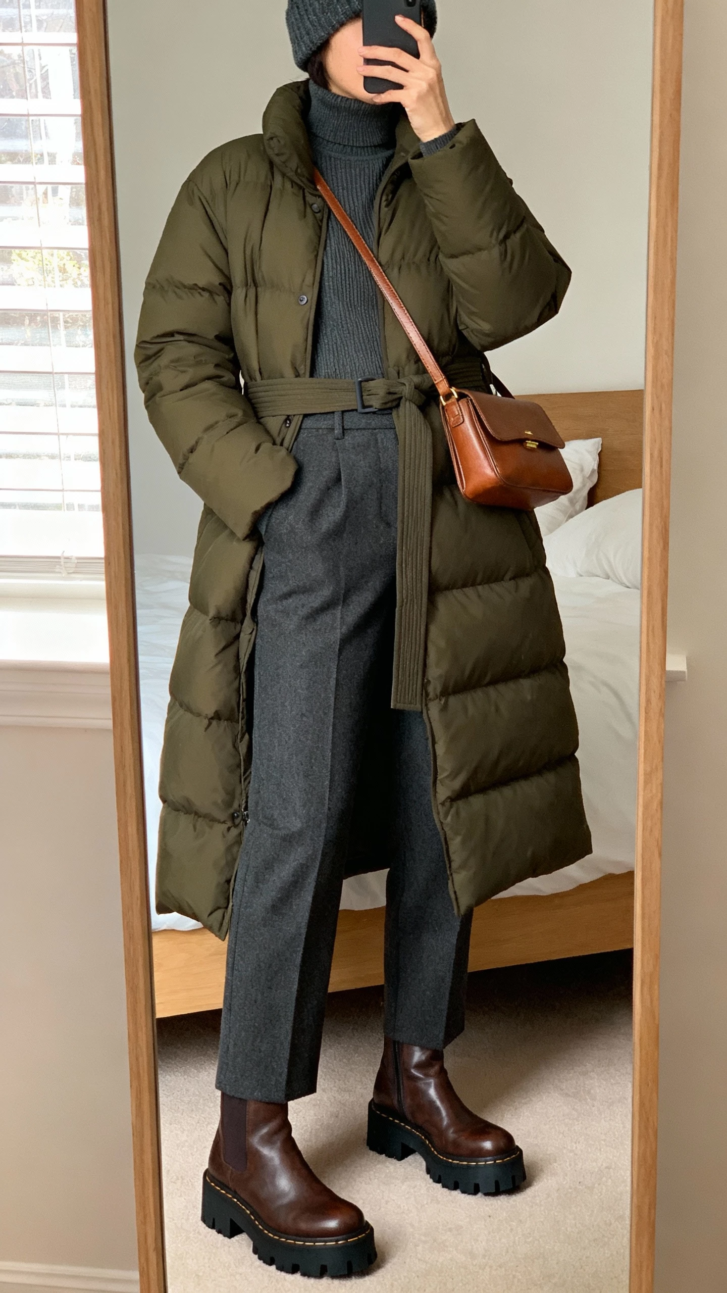 iPhone mirror selfie of a woman wearing a mid-length belted puffer coat in deep olive over a charcoal fine-gauge turtleneck, high-waisted tailored wool trousers, chunky-soled leather ankle boots, leather crossbody bag, and a wool beanie; face not visible, cropped at neck, bedroom full-length mirror, natural window light, natural iPhone photo quality.