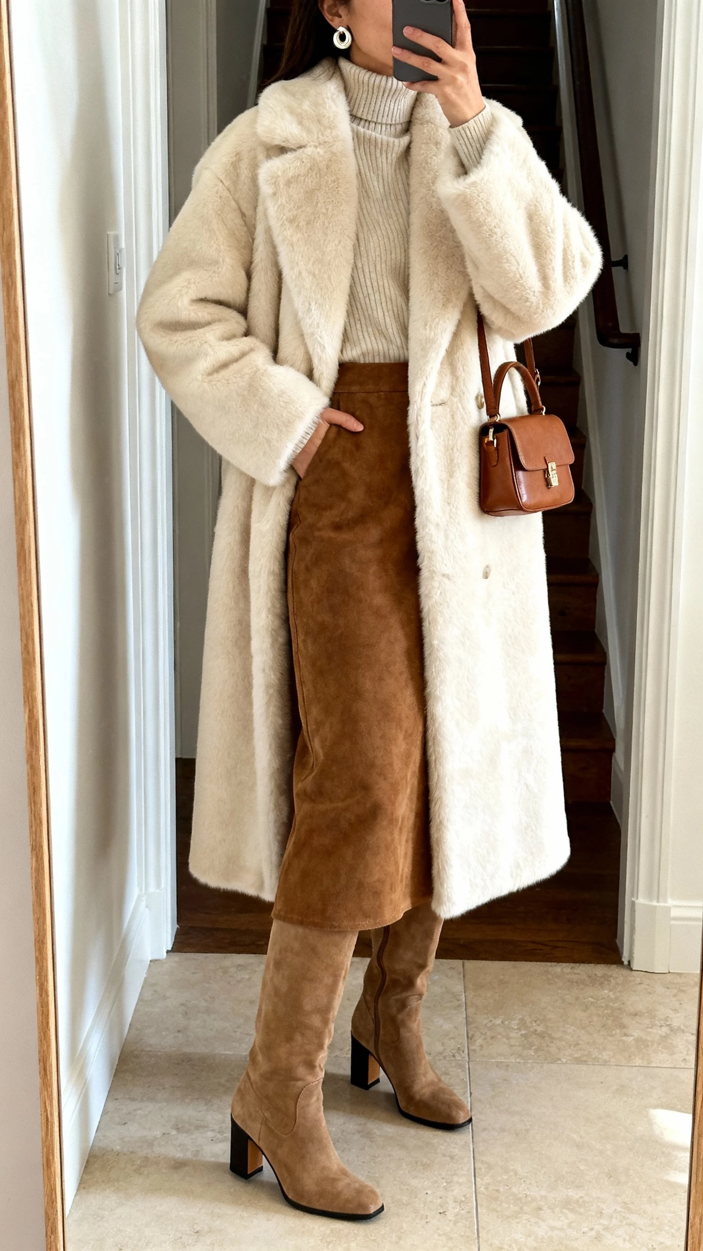 iPhone mirror selfie of a woman wearing a mid‑length cream faux‑fur coat, suede skirt, fine wool turtleneck, block‑heel knee‑high boots, leather mini bag, statement stud earrings; entryway mirror, face not visible, natural daylight, casual iPhone photo.