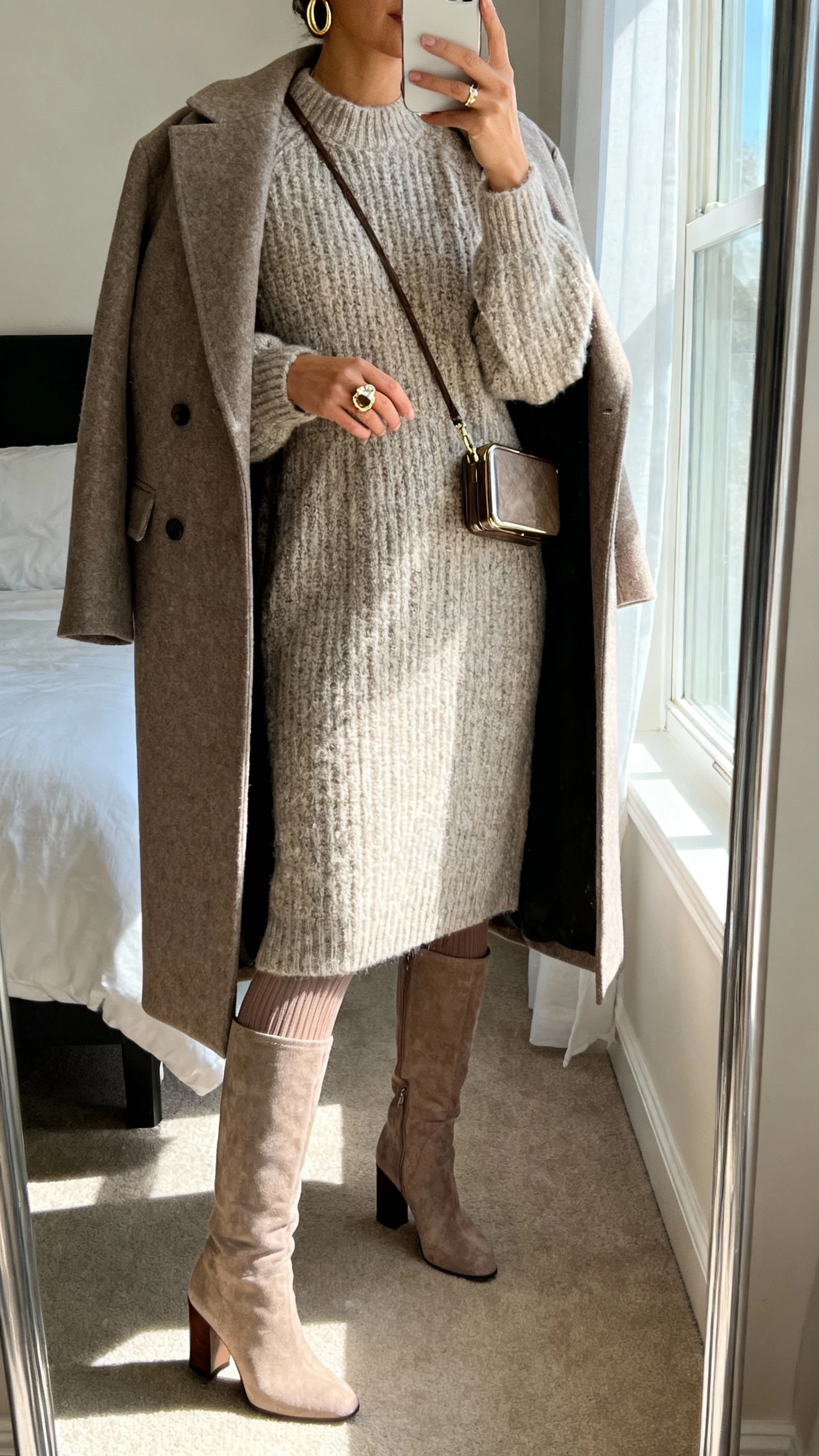 iPhone mirror selfie of a woman wearing a midi chunky sweater dress with knee‑high block‑heel boots, structured wool coat draped, opaque thermal tights, minimal gold hoops, statement ring, and a compact crossbody, face not visible, cropped at neck, bedroom mirror with window light, natural iPhone photo quality.