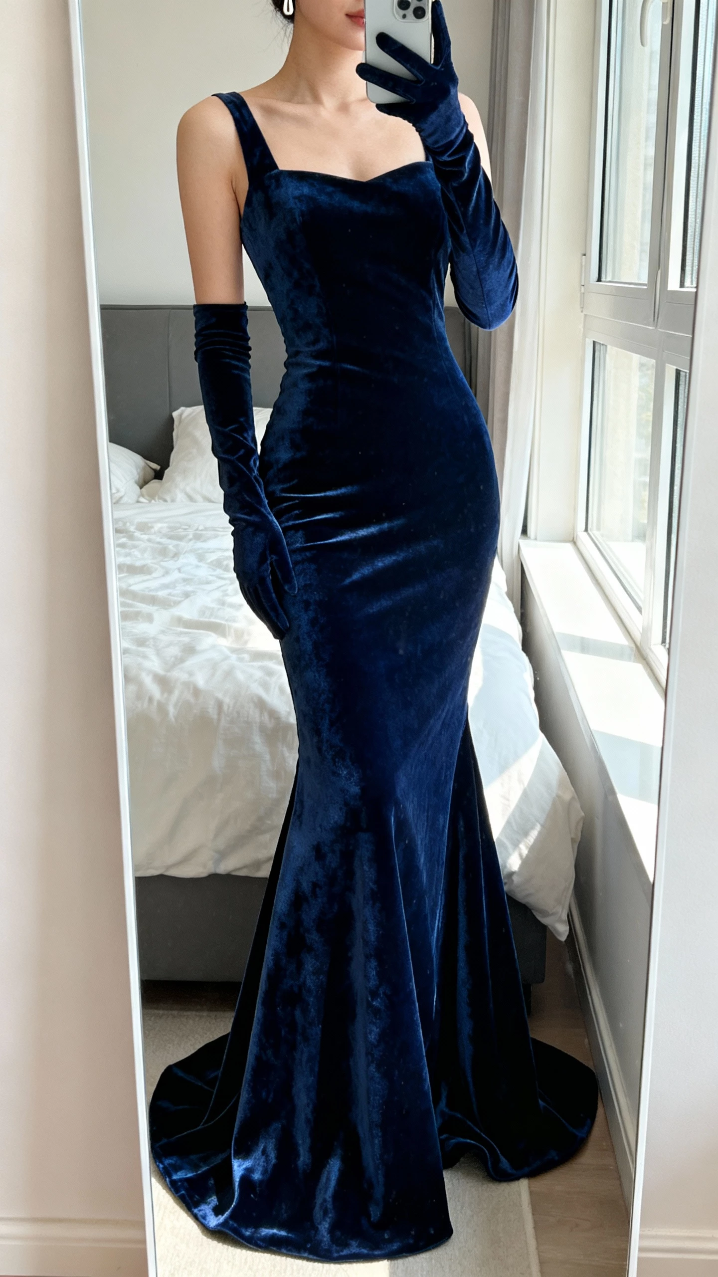 iPhone mirror selfie of a woman wearing a midnight blue velvet mermaid dress with built-in gloves and a dramatic flared hem, minimal earrings, face not visible, cropped at neck, bedroom mirror with soft window light, natural iPhone photo quality.