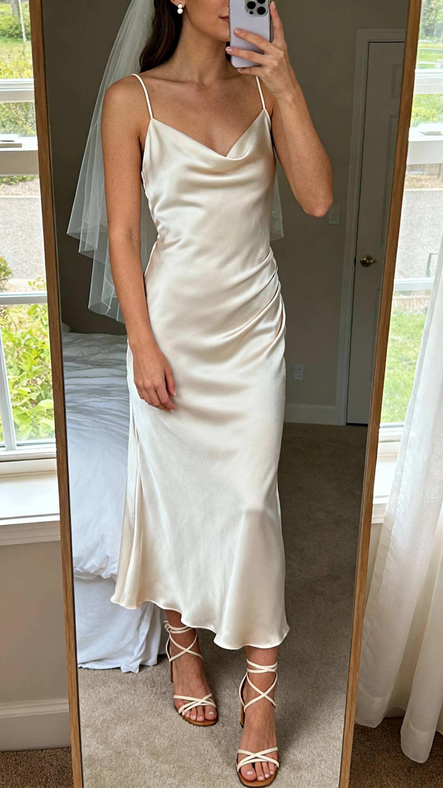 iPhone mirror selfie of a woman wearing a minimalist silk slip wedding dress with bias-cut drape, delicate spaghetti straps, simple veil, dainty stud earrings, and strappy heeled sandals, face not visible, cropped at neck, bedroom mirror with soft window light, natural iPhone photo quality.