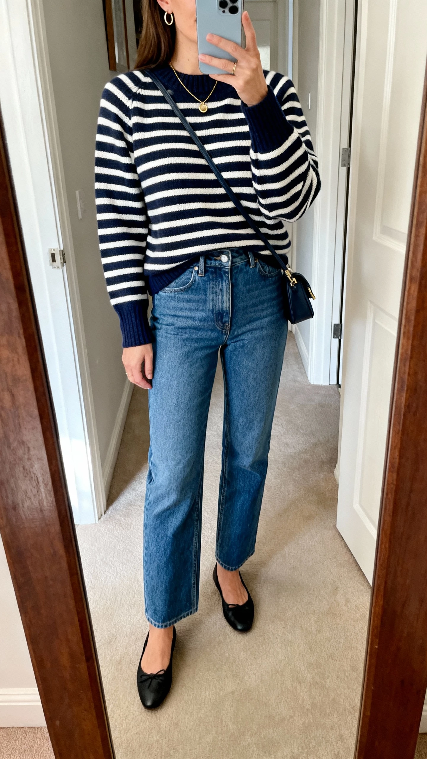 iPhone mirror selfie of a woman wearing a navy-and-white striped Breton sweater, high-rise straight blue jeans, black ballet flats, small crossbody, delicate gold necklace, thin hoops; entryway mirror, face not visible, soft morning light, natural iPhone photo quality.