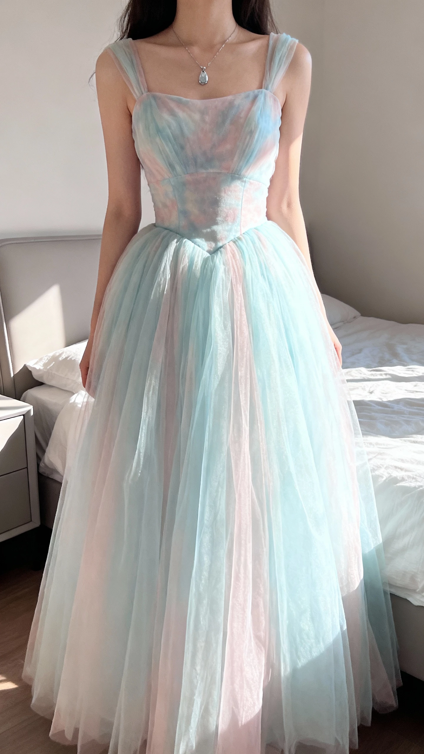 iPhone mirror selfie of a woman wearing a pastel ball gown in soft blush/ice-blue tones with airy tulle skirt, subtle color-wash bodice, and delicate pendant necklace, face not visible, cropped at shoulders, sunlit bedroom, natural iPhone photo