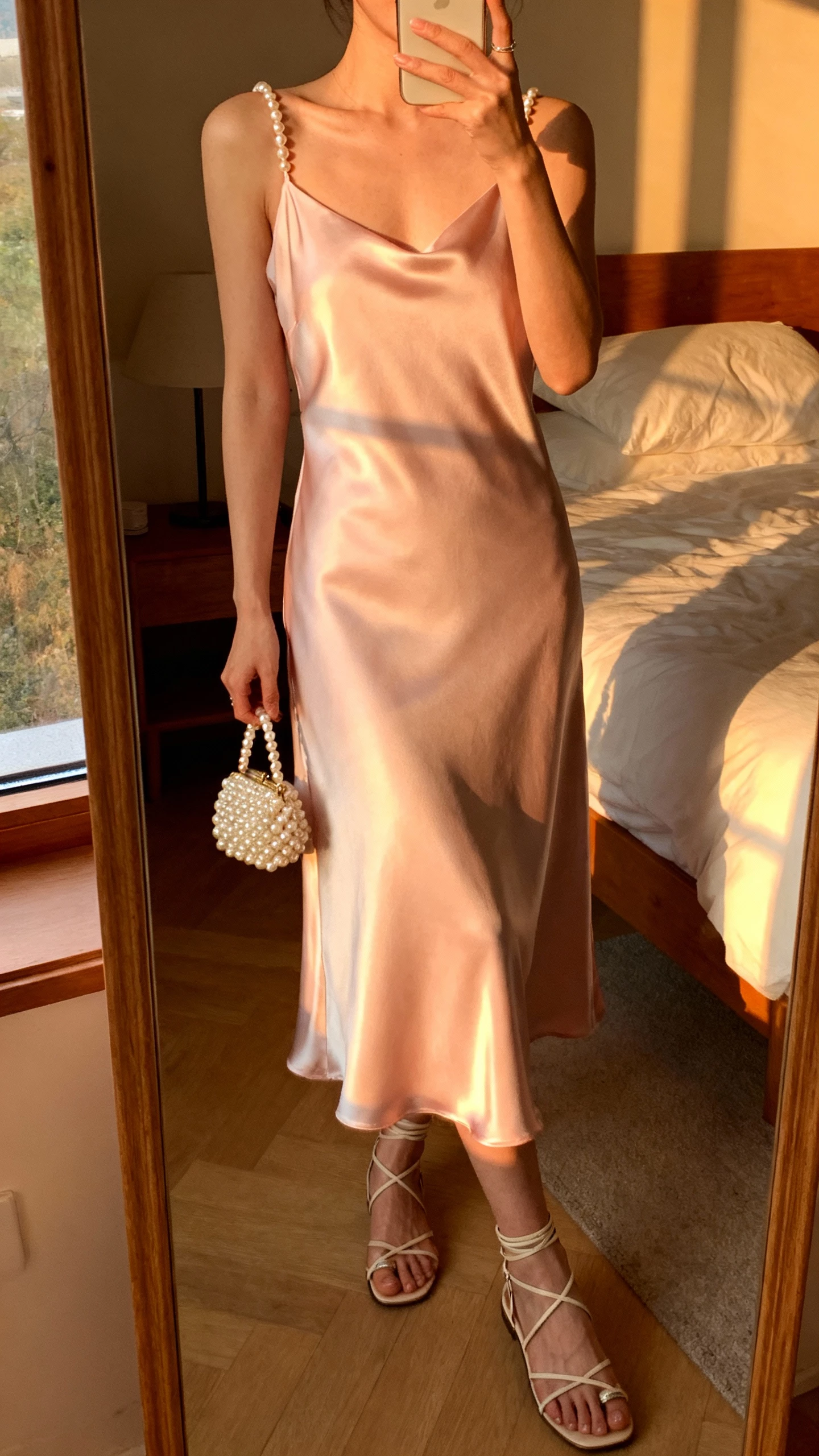 iPhone mirror selfie of a woman wearing a pastel satin slip dress with delicate pearl straps and a small pearl clutch, strappy sandals, face not visible, cropped at neck, bedroom mirror at golden hour with warm window light, natural iPhone photo quality.