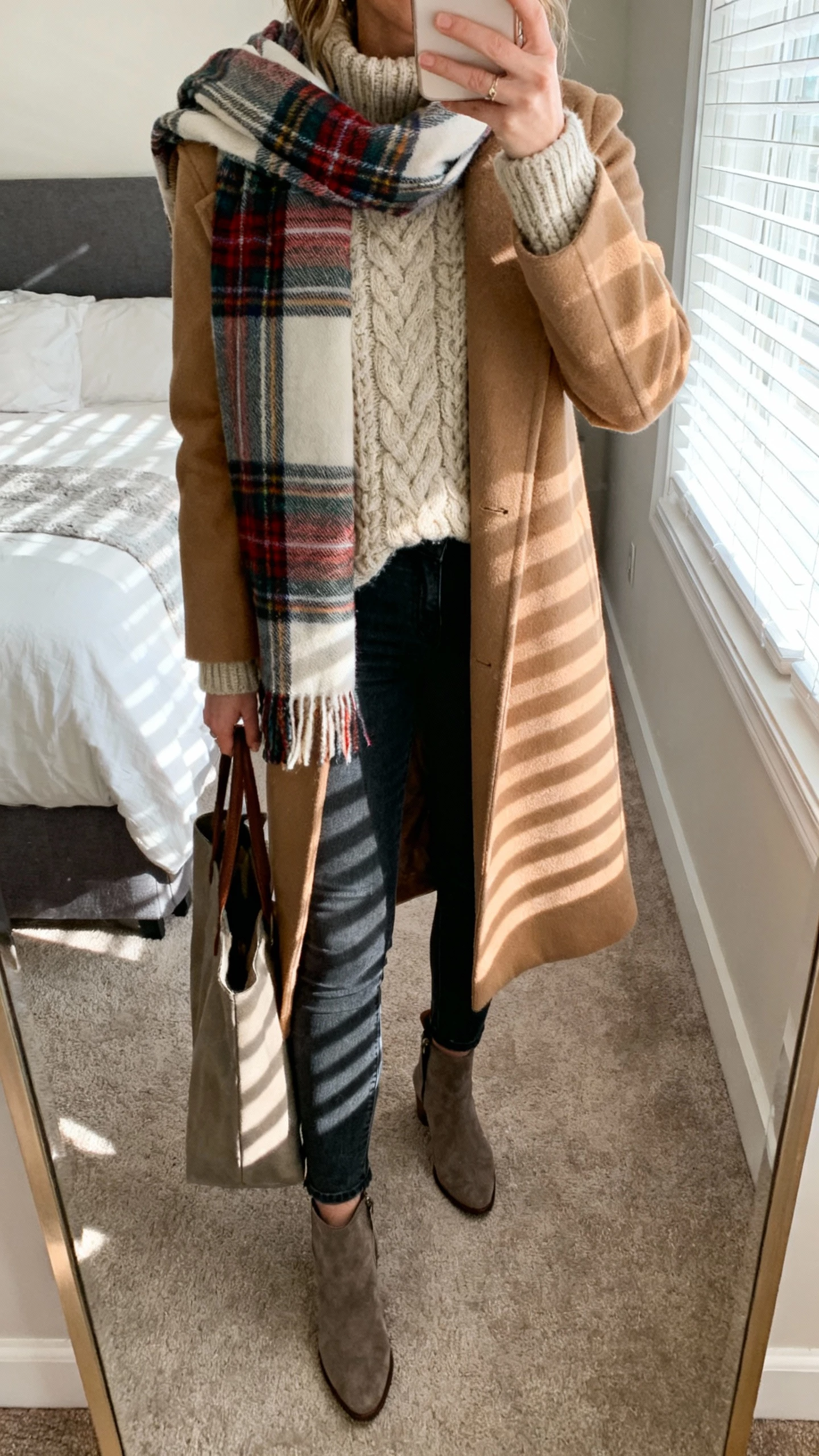 iPhone mirror selfie of a woman wearing a plaid scarf over a camel coat, chunky knit sweater, dark skinny jeans, and ankle boots, holding a tote, face not visible, cropped at neck, bedroom mirror with morning window light, natural iPhone photo quality.