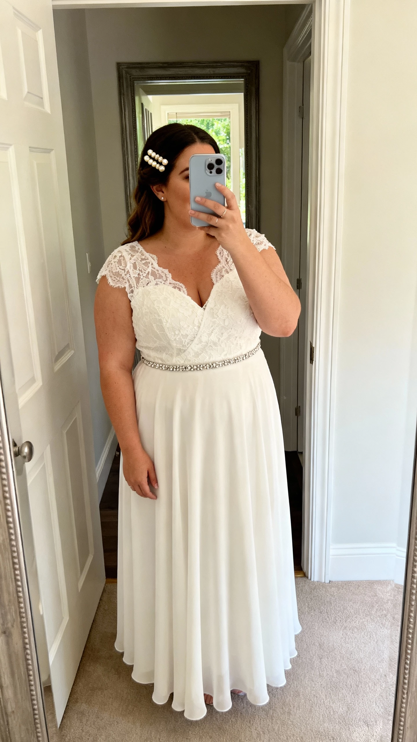 iPhone mirror selfie of a woman wearing a Rebecca Ingram Curve lightweight chiffon A-line with soft lace bodice, V-neck, and slim beaded belt, paired with pearl hairpins; face not visible, entryway mirror, afternoon natural light, casual iPhone snap.