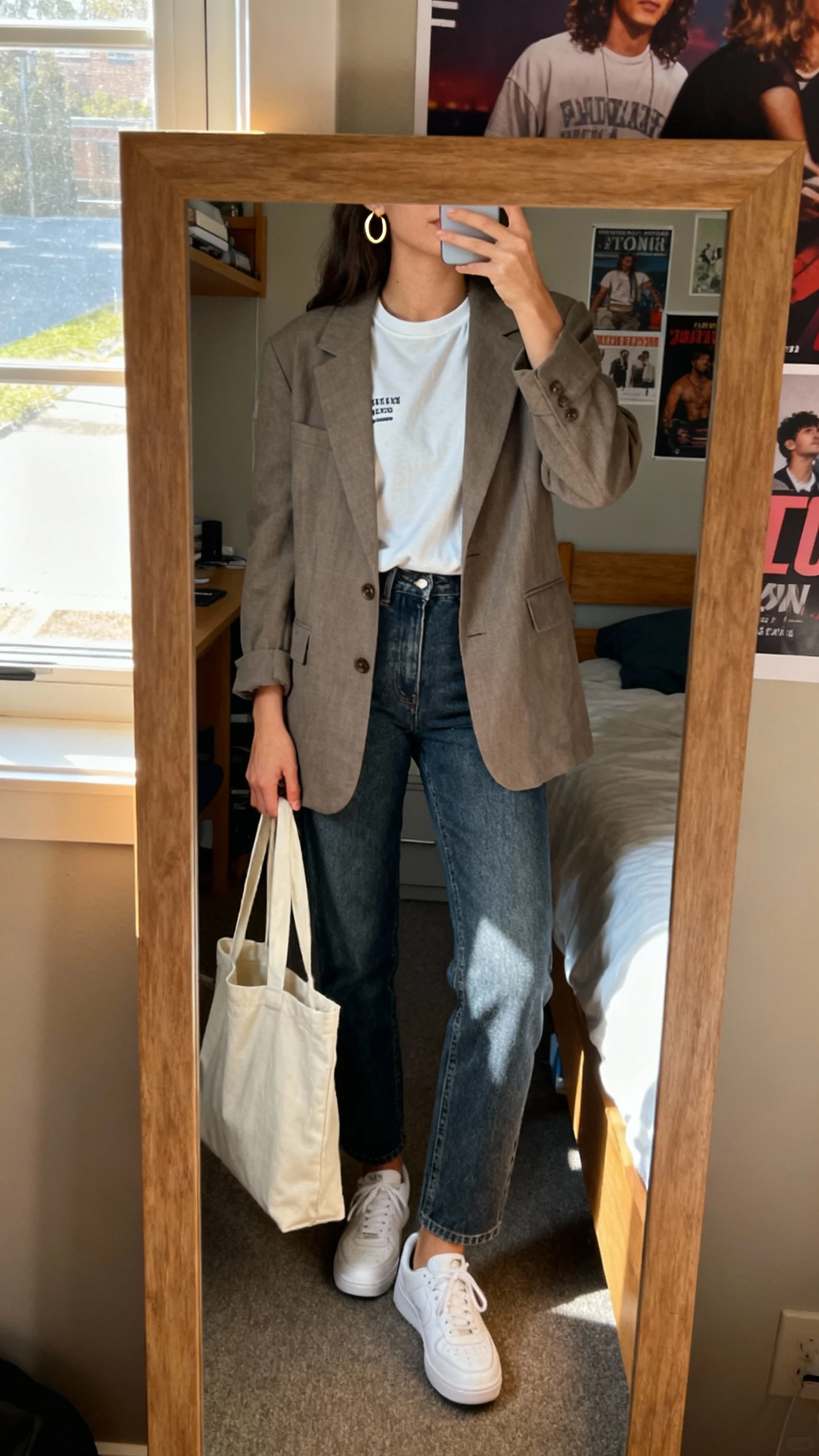 iPhone mirror selfie of a woman wearing a relaxed blazer over a classic crewneck tee, straight-leg jeans, white sneakers, simple gold hoop earrings, tote bag in hand, face not visible, cropped at neck, dorm bedroom mirror with posters, morning window light, natural iPhone photo quality