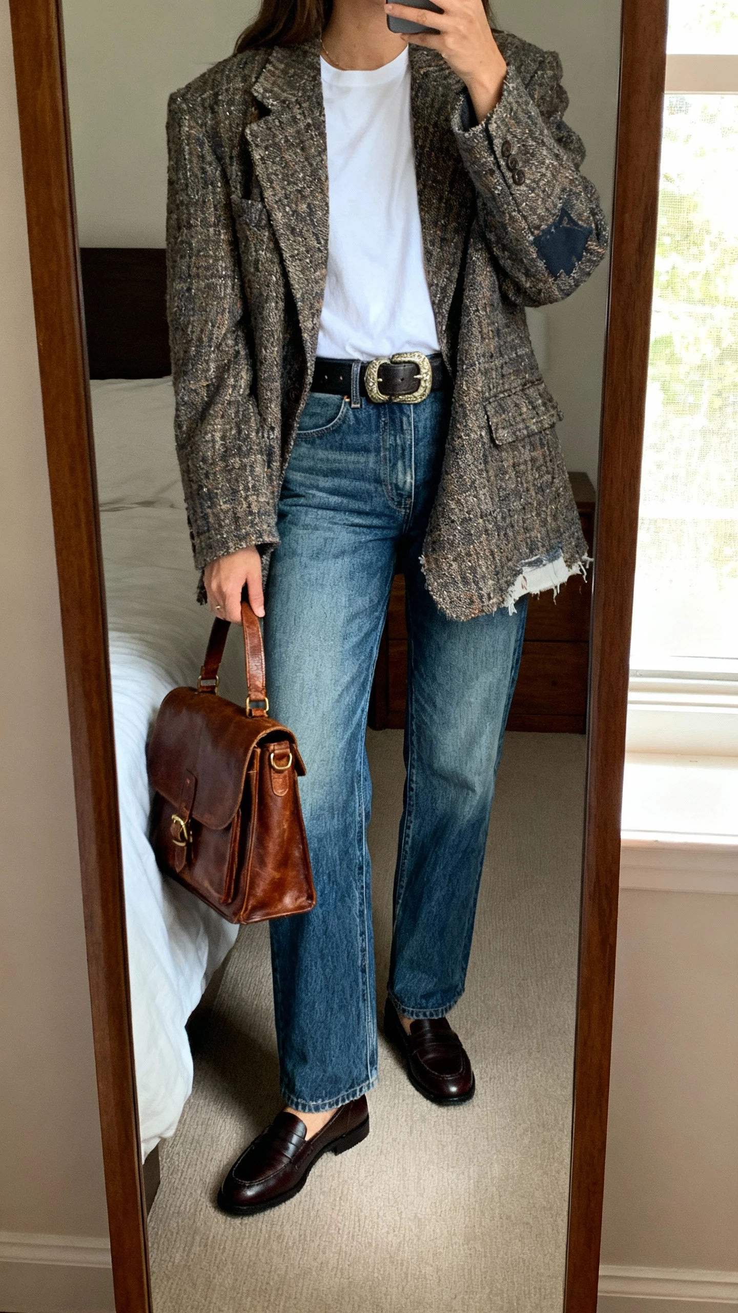 iPhone mirror selfie of a woman wearing a reworked oversized tweed blazer over a crisp white tee, straight-leg vintage blue denim, chunky belt, loafers, and a thrifted leather satchel, face not visible, cropped at neck, bedroom mirror with natural window light, natural iPhone photo quality.