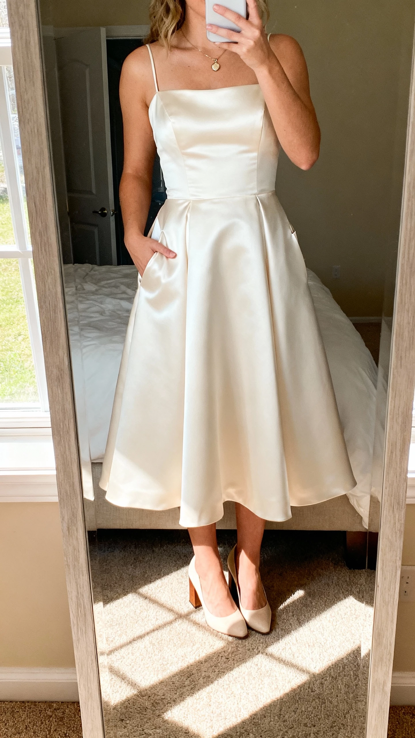 iPhone mirror selfie of a woman wearing a satin A-line wedding dress with pockets, clean skirt, subtle sheen, simple pendant, and block heels, hands in pockets; face not visible, bedroom mirror, bright morning window light, casual iPhone aesthetic.